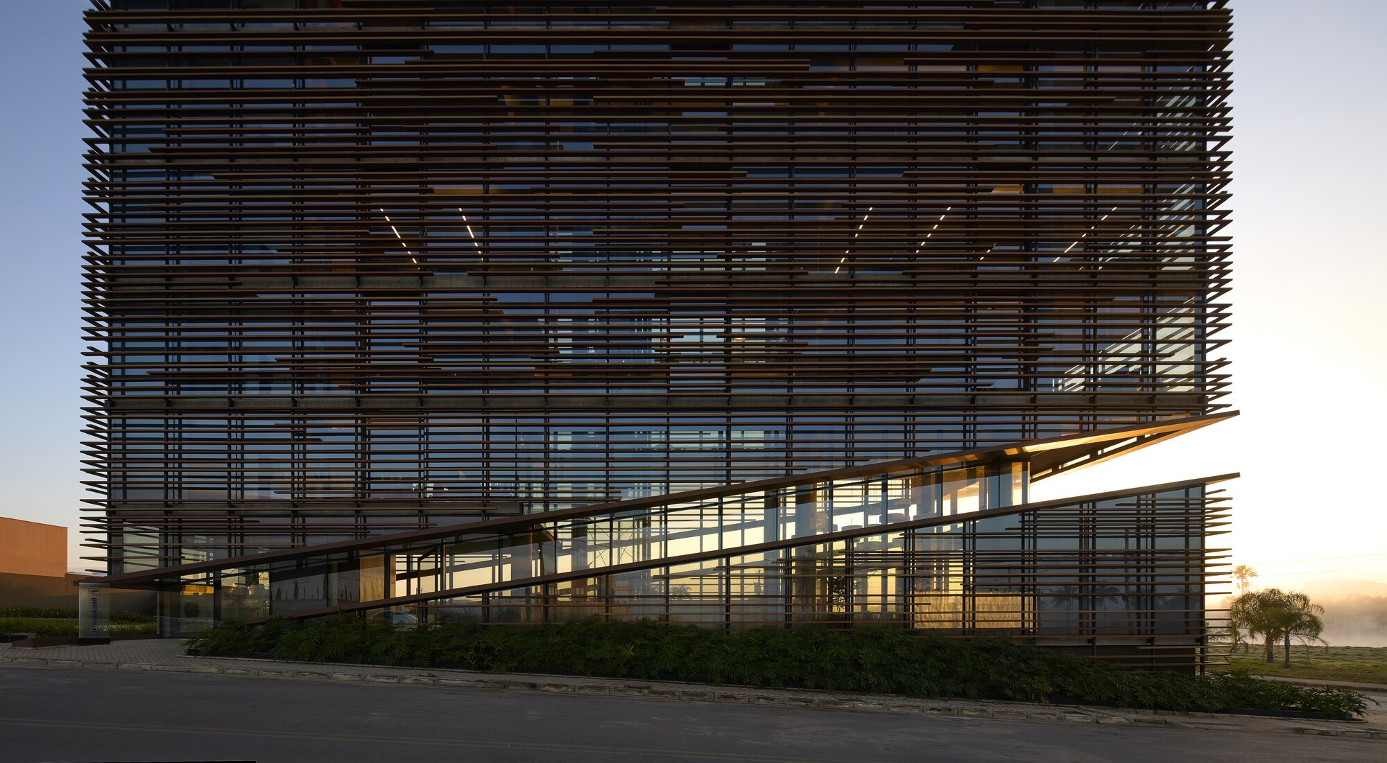 Ground-level facade showing the angled glazed base cut beneath the louvered block, with a shrub hedge and morning mist at right