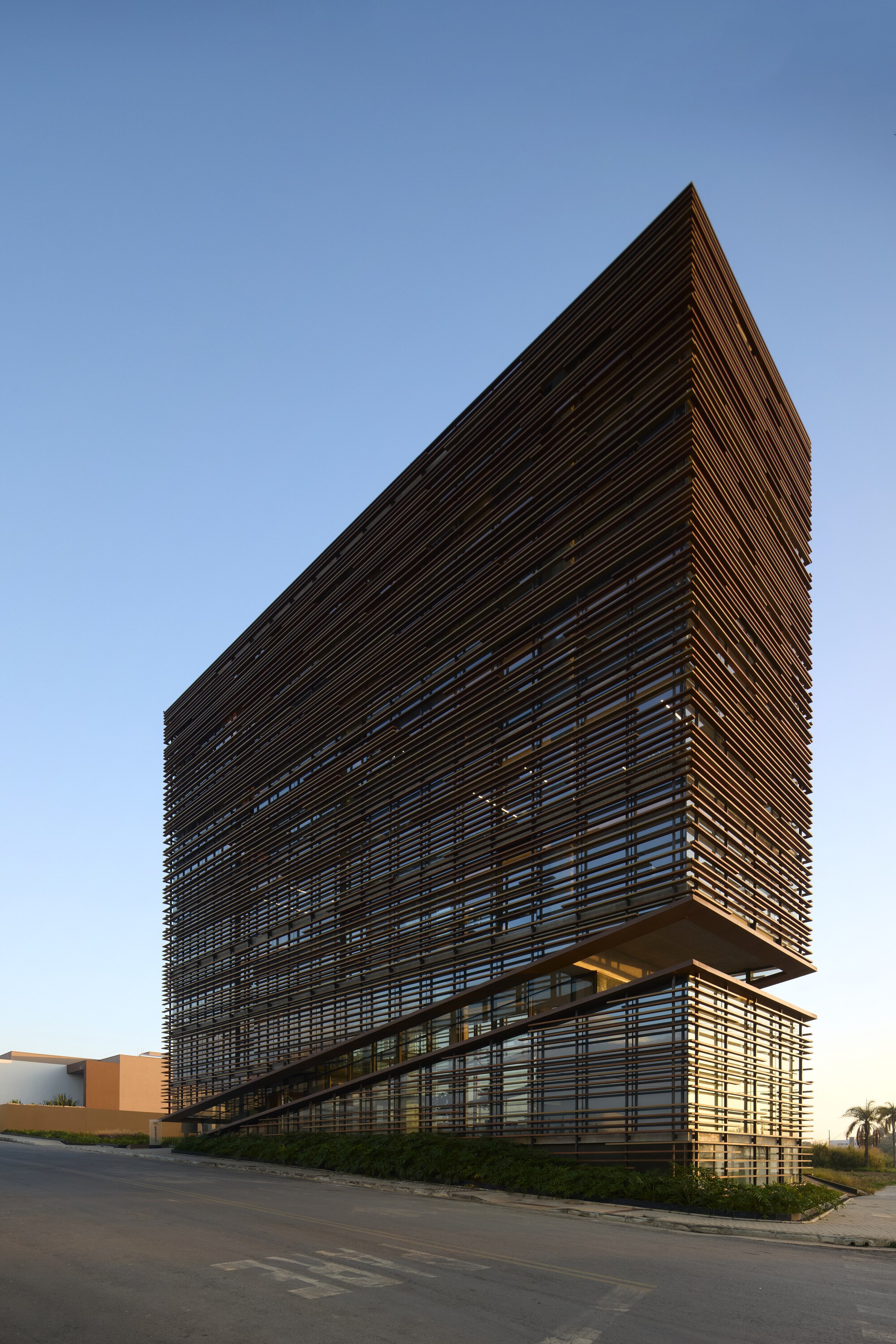 Corner of the tall timber-louvered office block with a tilted base volume, seen from the street under a clear evening sky