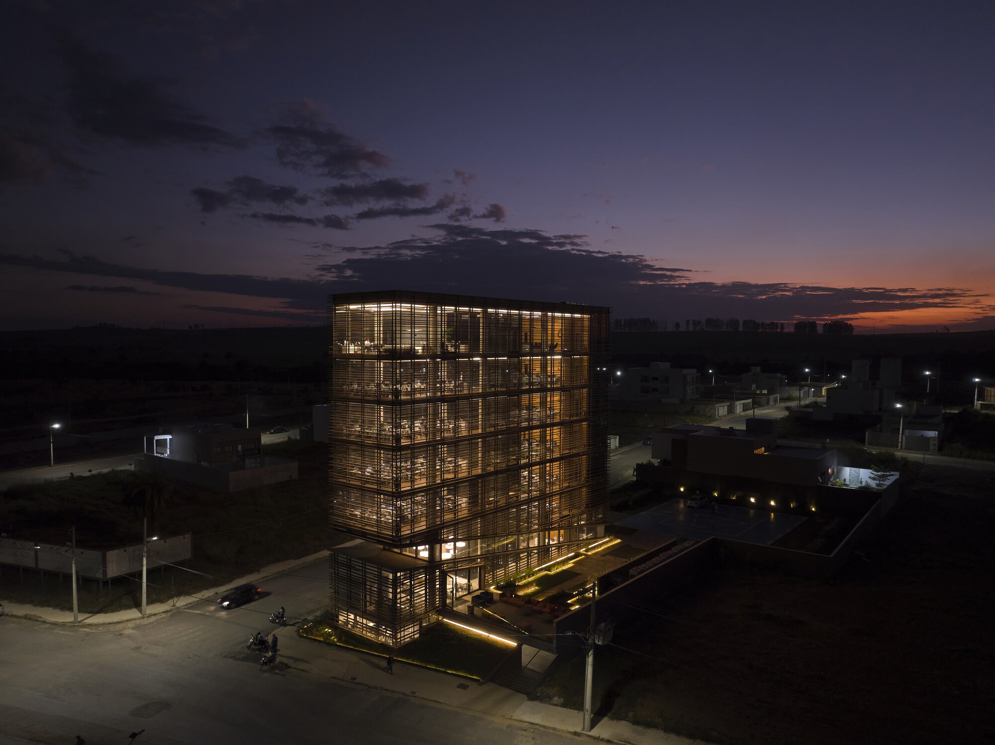 Aerial view of the glowing louvered tower at dusk, with lit entrance terrace and a band of orange sky on the horizon