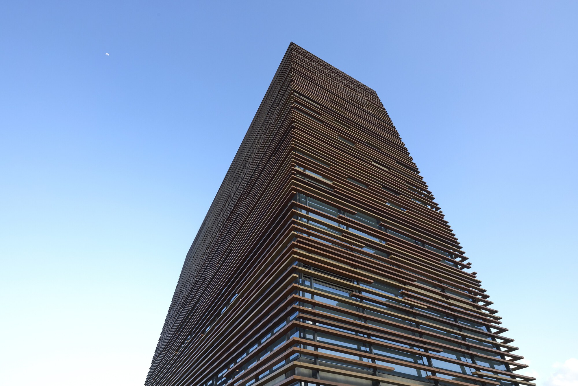 Upward view of the horizontal timber slats wrapping the tower, with a faint moon in the clear blue sky