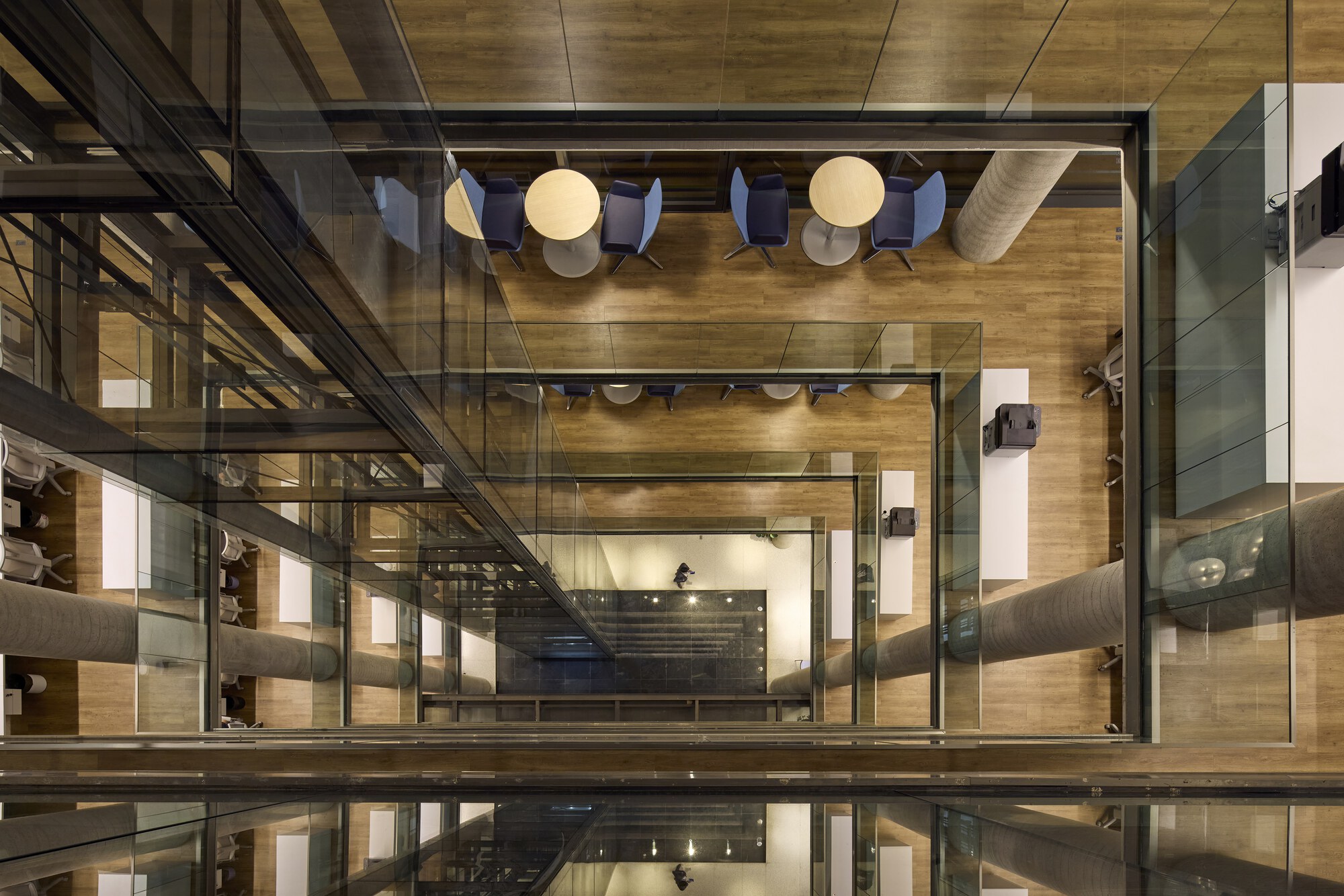 Looking straight down the glass-edged atrium, past stacked floors with round tables, blue chairs and large pale ducts, under artificial light
