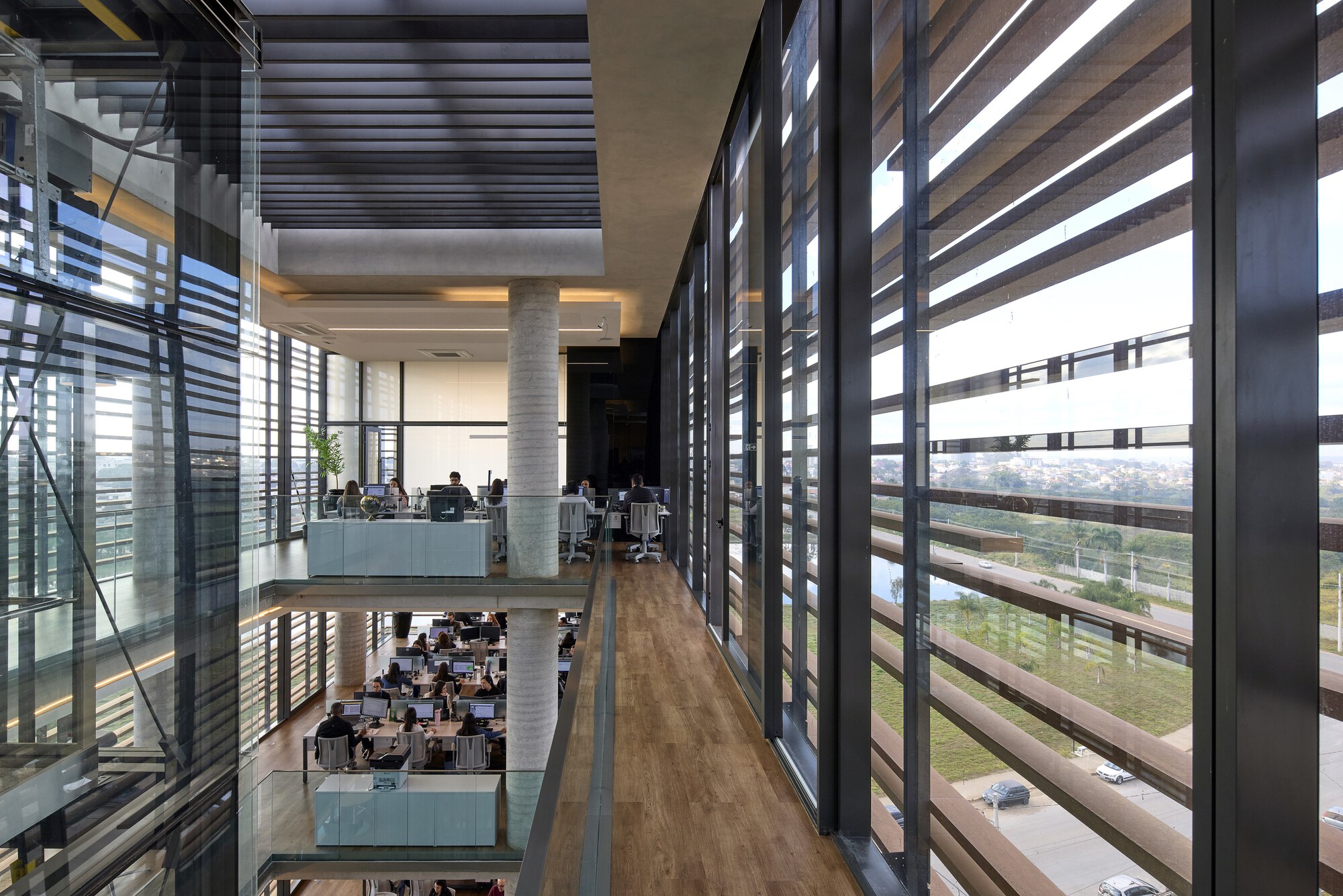 Timber-floored walkway beside the louvered glass wall, overlooking open-plan desks, concrete columns and a road with parked cars in daylight