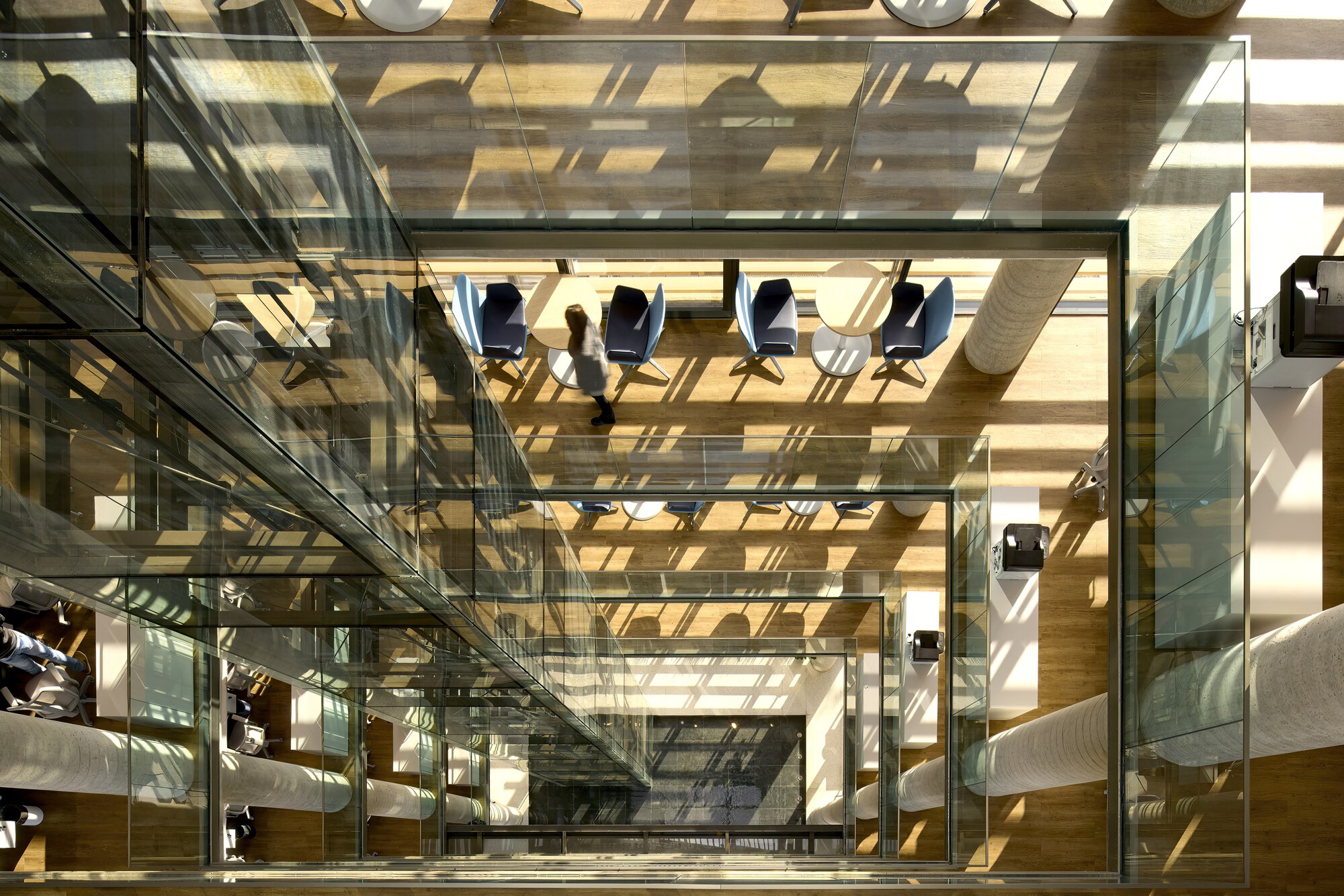 Top-down view of the glass atrium with a person crossing the lounge floor, sunlight casting slatted shadows across chairs and ducts