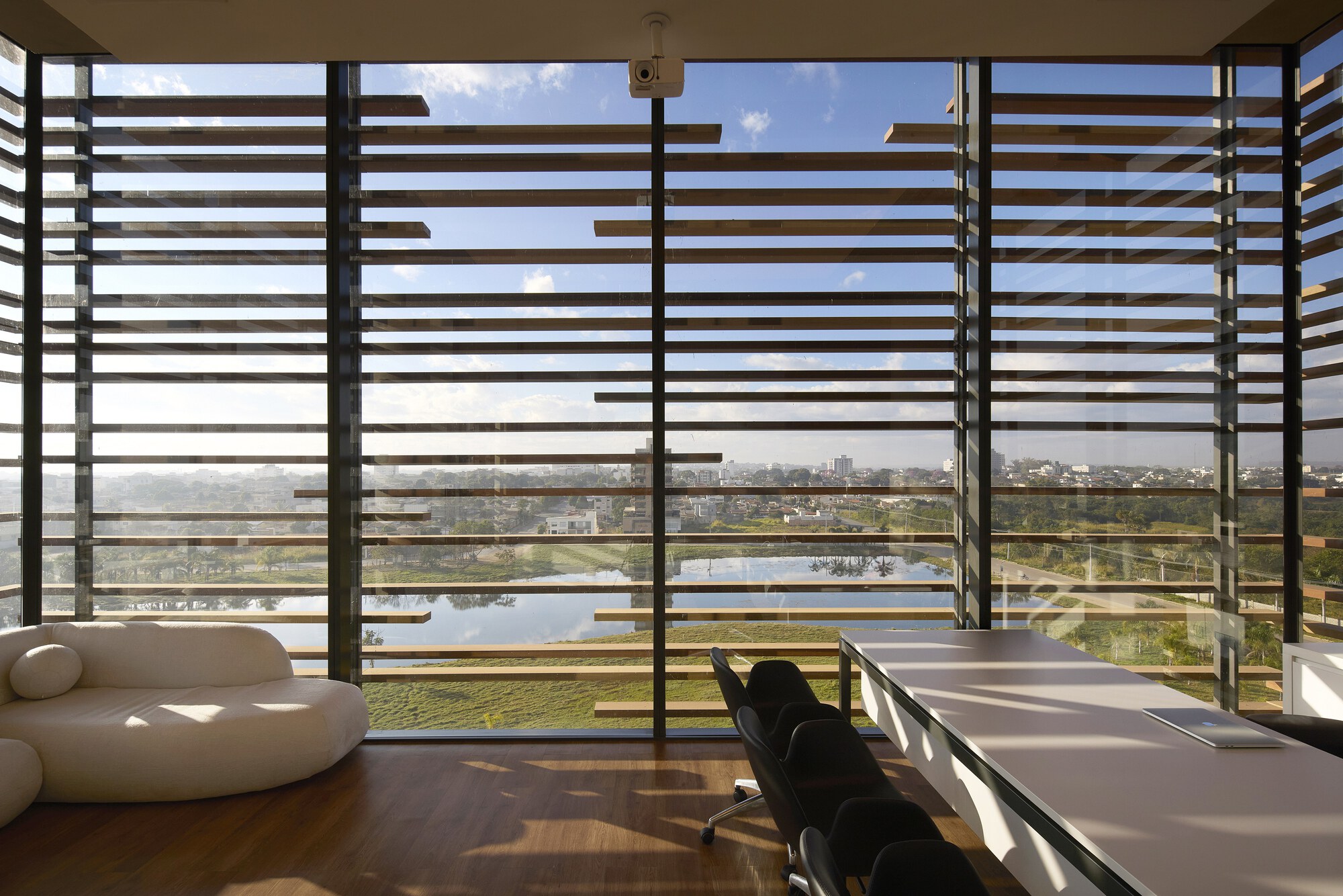 Office corner with floor-to-ceiling glass and timber louvers framing a lake view, with a curved white sofa and long table in afternoon sun