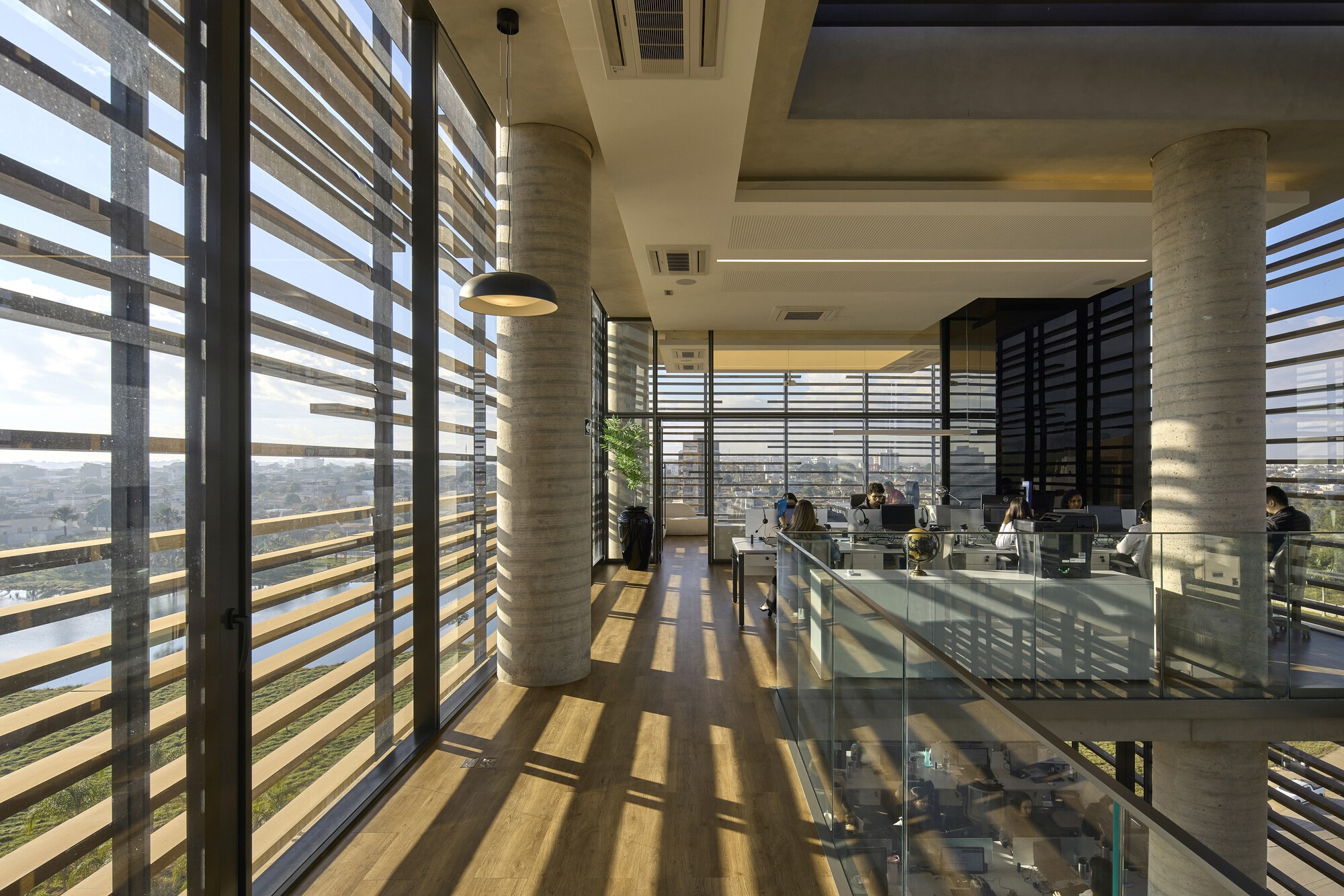 Upper-floor walkway beside louvered glazing, concrete columns and a glass balustrade overlooking staff at desks, with sunlight striping the timber floor
