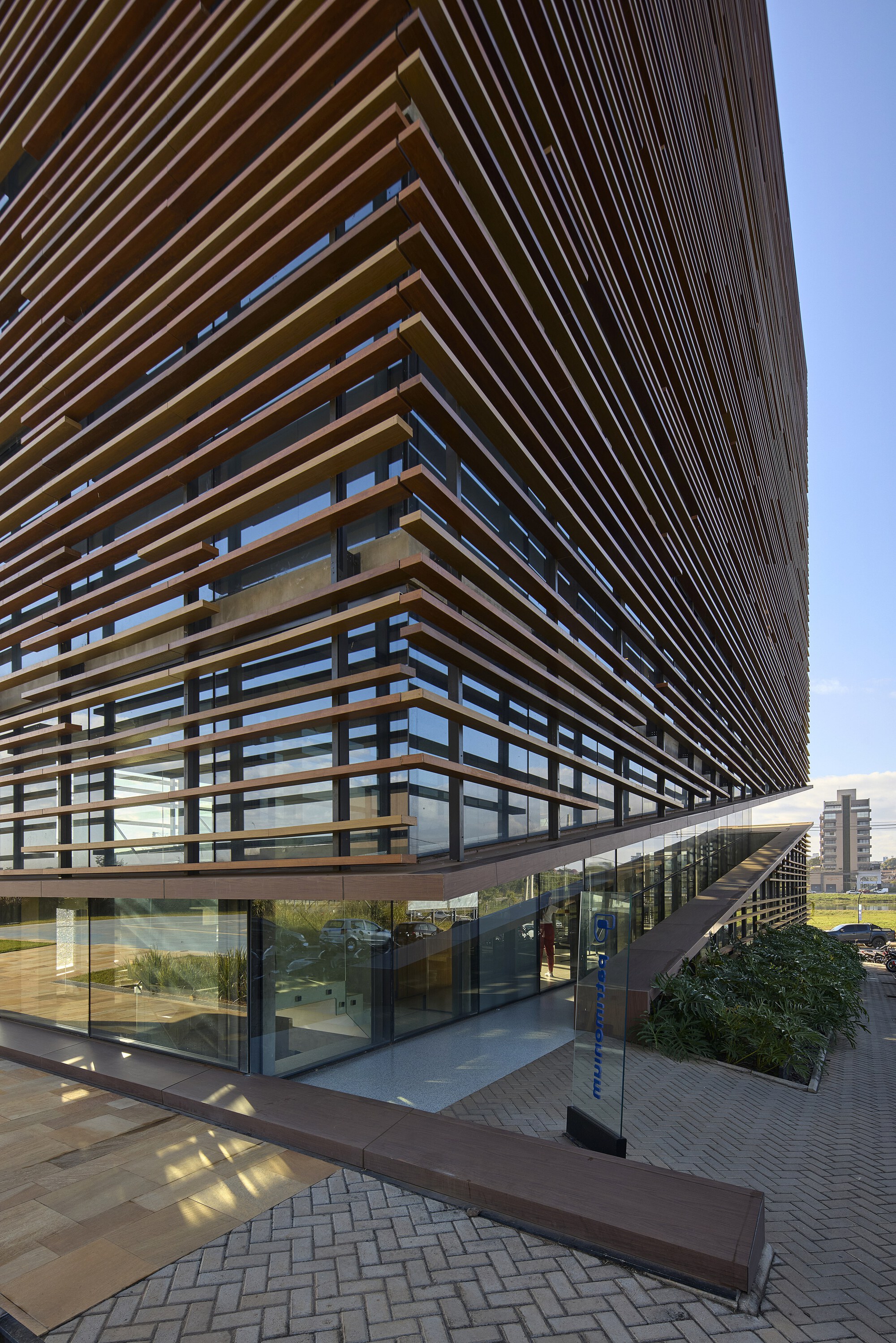 Corner of the timber-louvered facade rising above a glazed entrance, with a planted bed and herringbone brick paving under clear sky