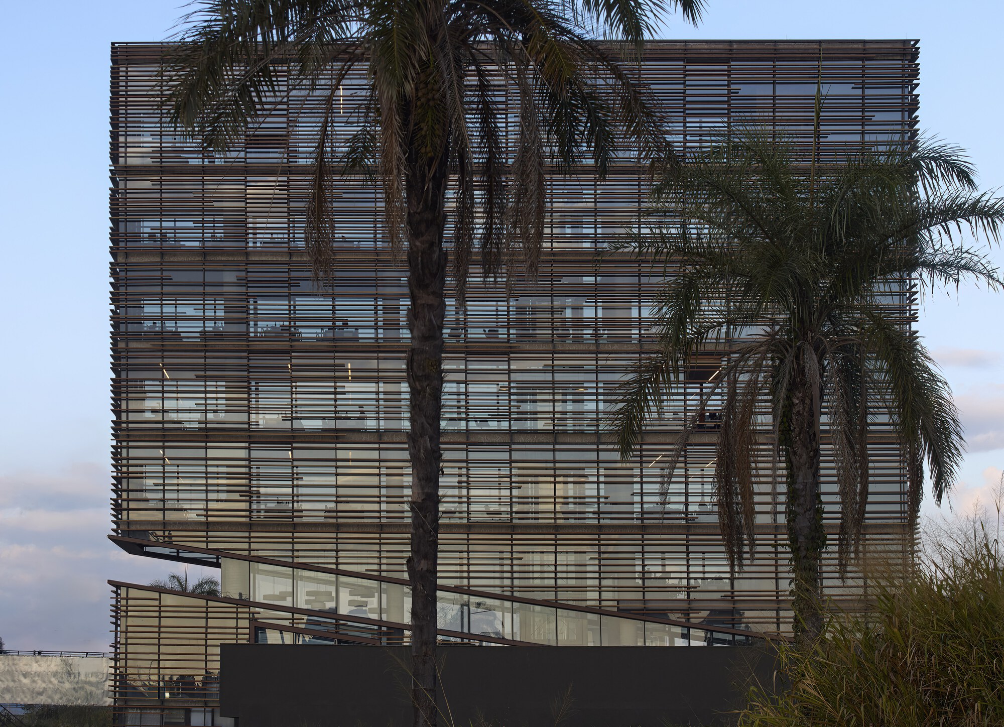 Square louvered facade seen through two palm trees, with a sloped ramp at its base under a pale evening sky