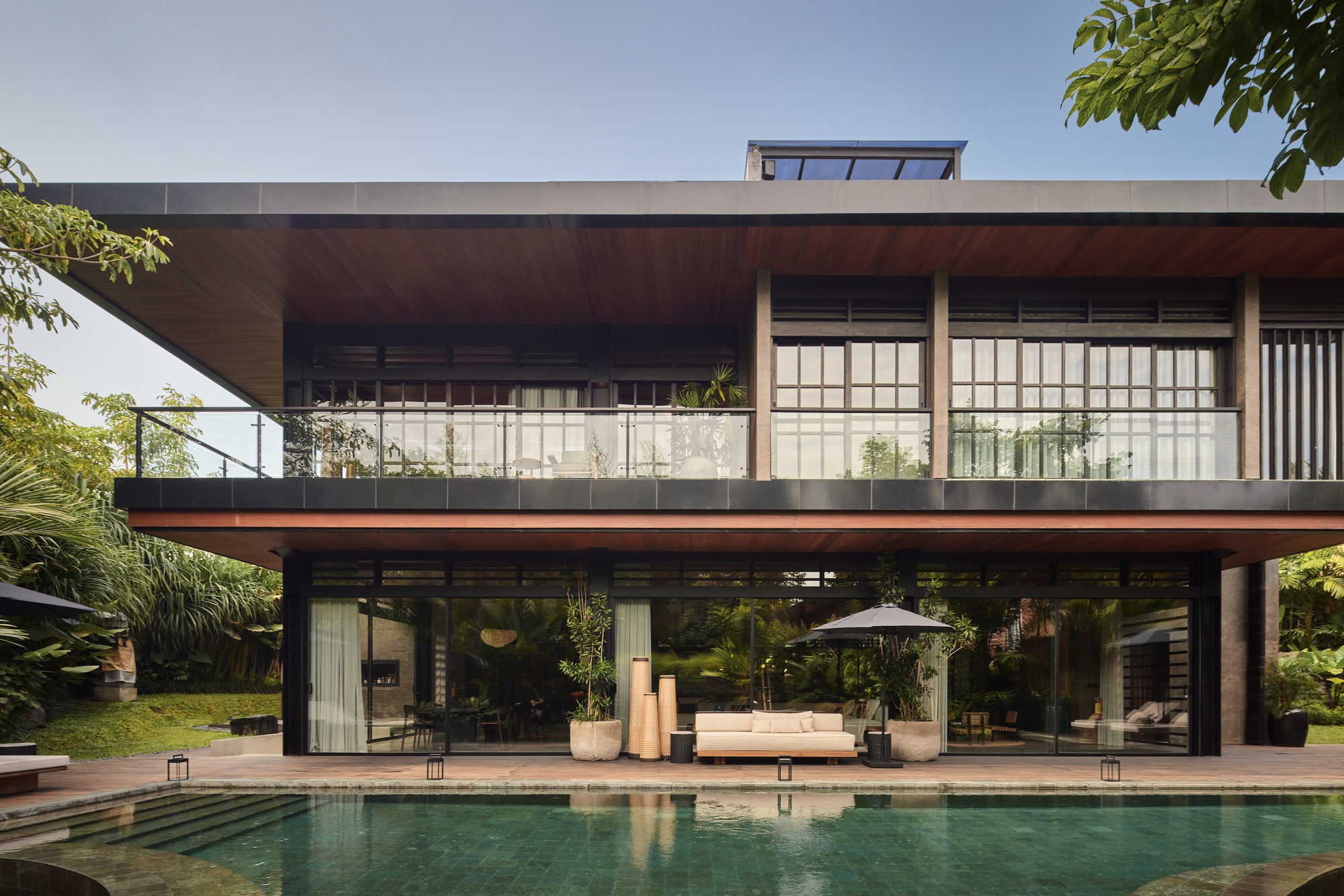 Two-storey facade with deep timber-lined overhangs and glass balustrade rising behind a green-tiled pool in clear daylight