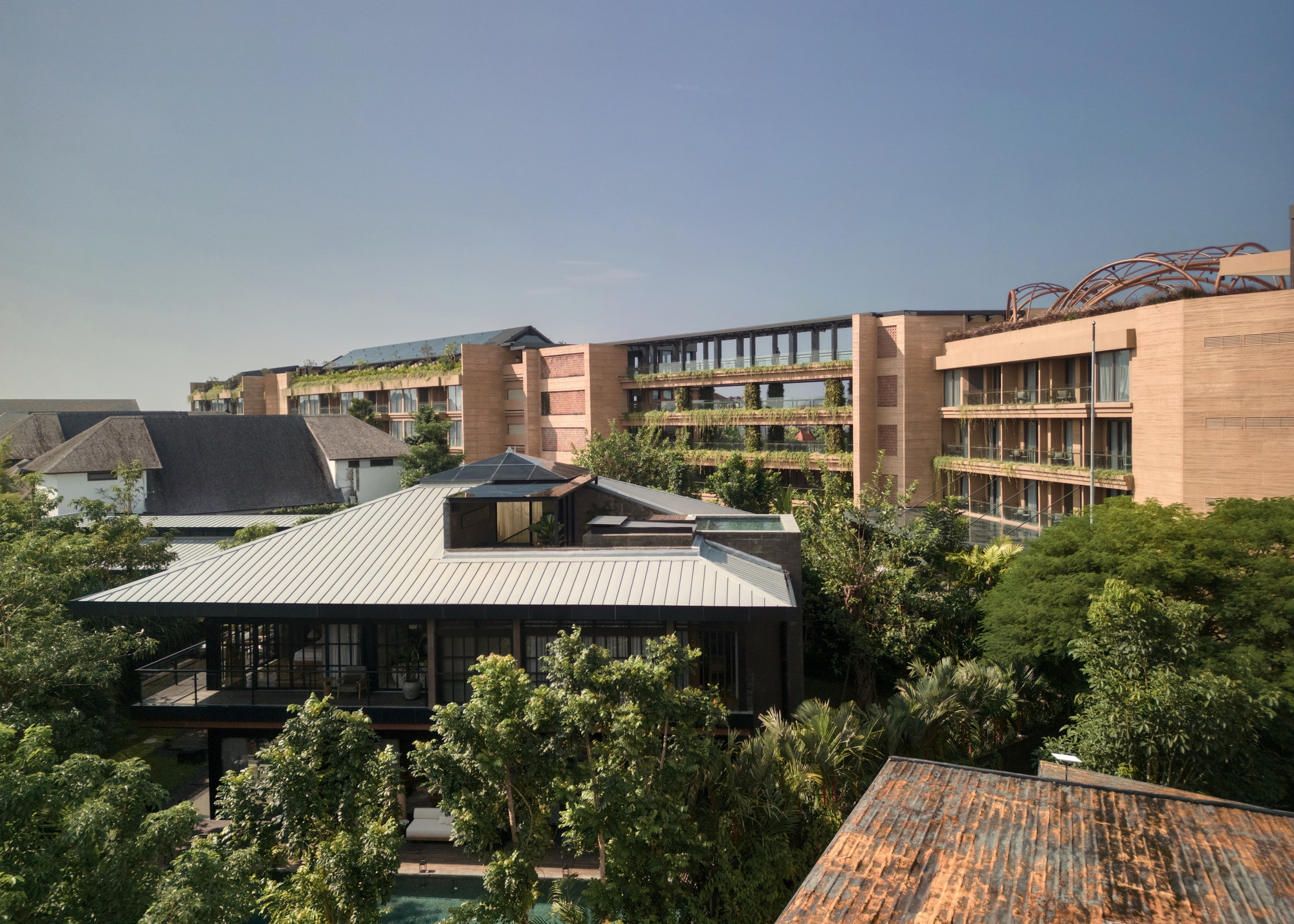 Aerial view of a metal-roofed pavilion in front of terraced blocks with planted balconies, seen among treetops in warm afternoon light