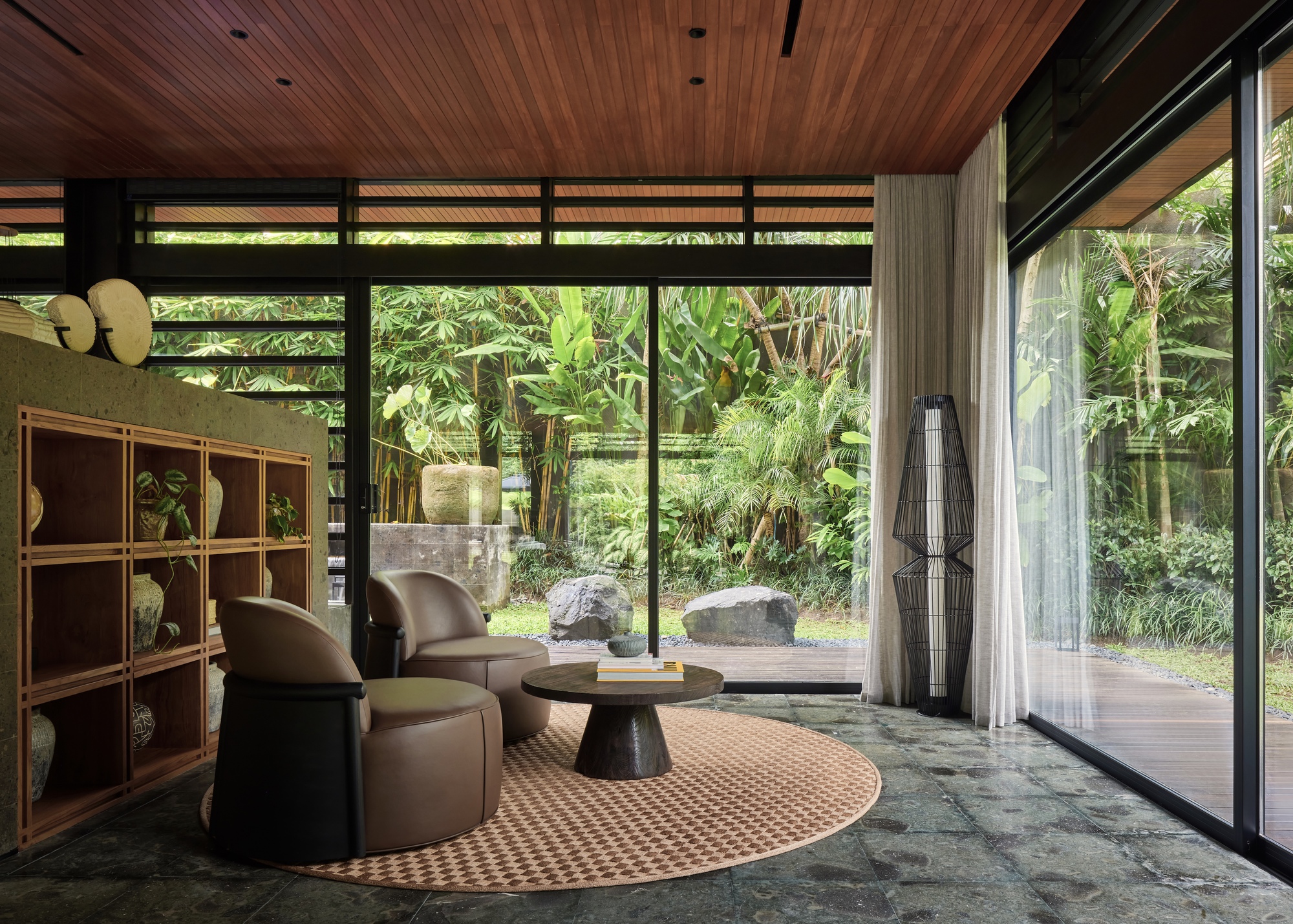 Living corner with round woven rug, leather armchairs and stone-clad shelving facing a bamboo garden through floor-to-ceiling glass