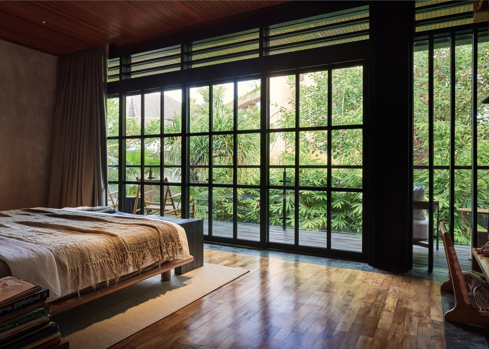 Bedroom with black steel-framed glazing opening to a timber deck and dense foliage, light falling across the teak floor