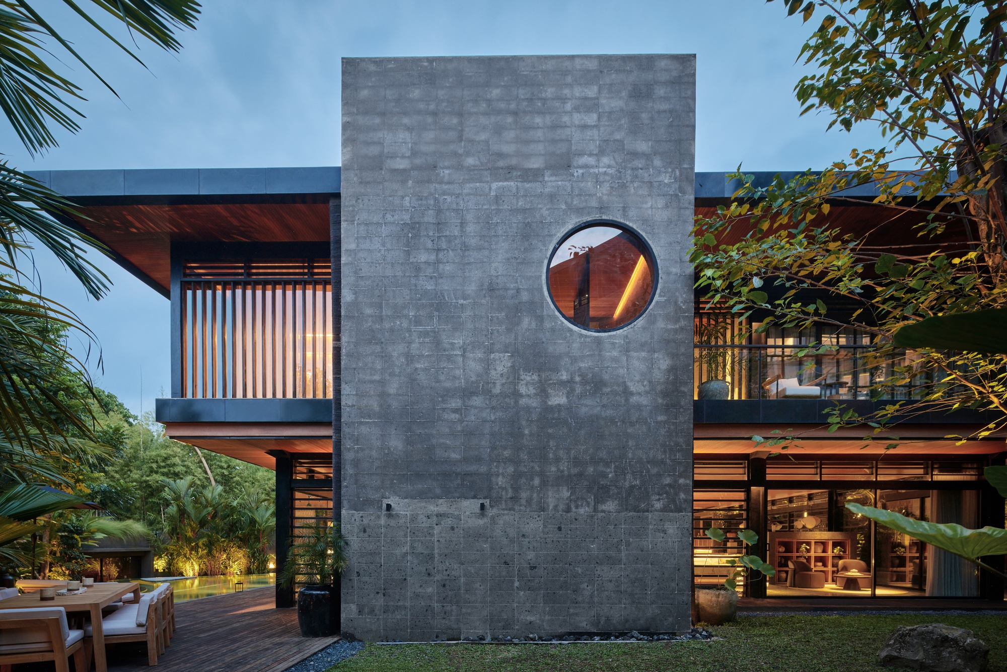Board-formed concrete wall with a circular window stands between lit timber-screened rooms at dusk, with a lawn and palms around it
