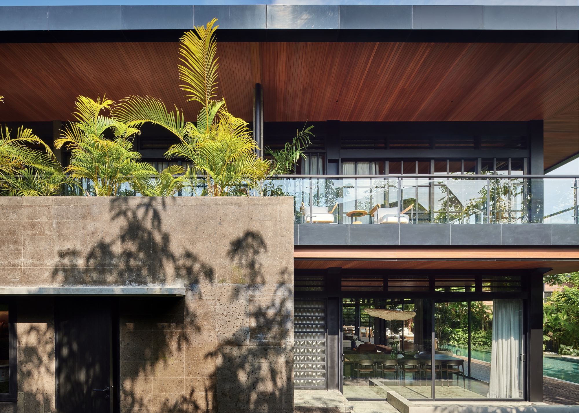 Rough volcanic stone wall with a planted palm terrace above, casting leaf shadows beside a glazed dining room and pool
