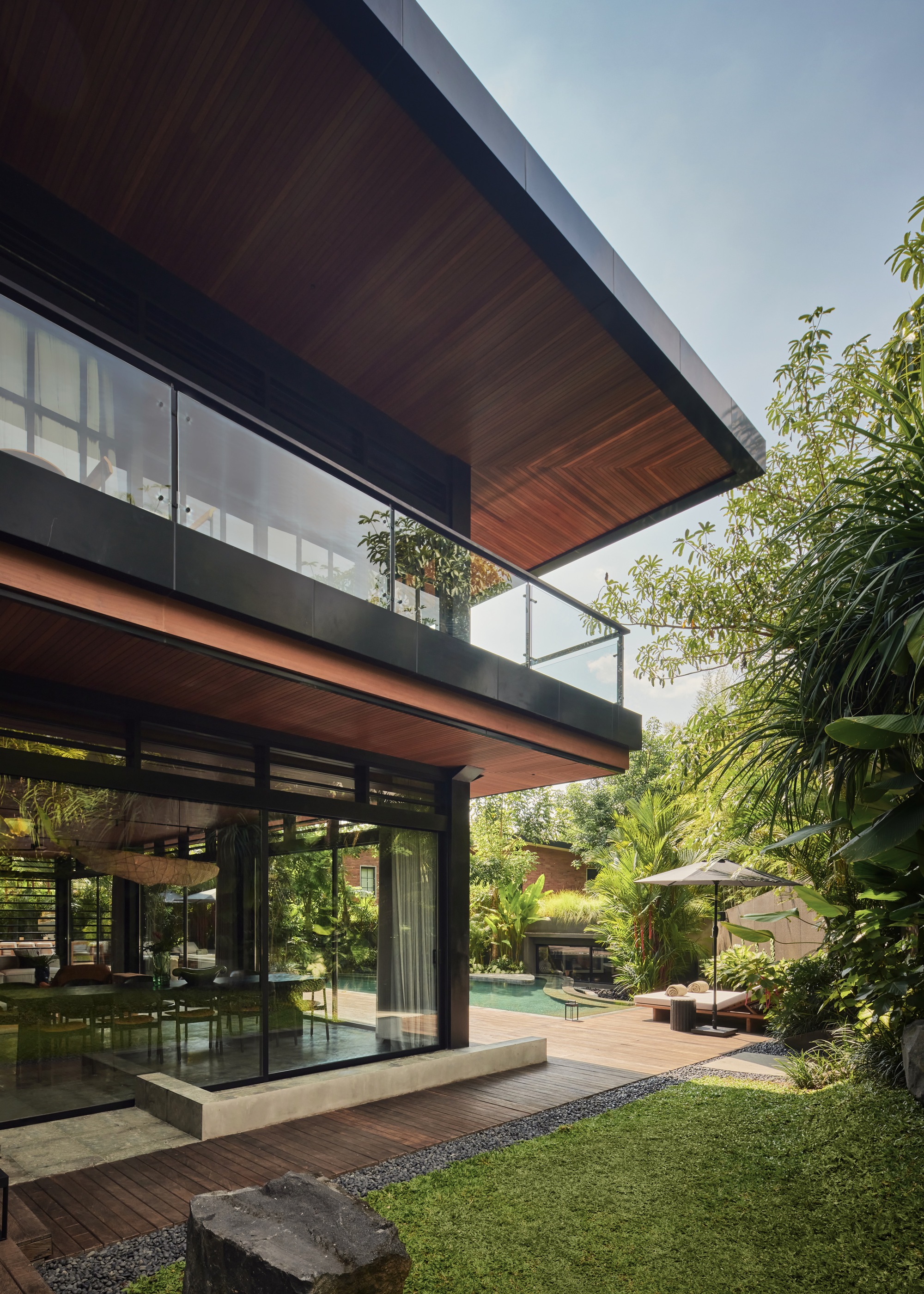 Two-storey timber-soffit volume with glass balustrade overlooking a lawn, wood deck and pool, framed by tropical foliage in afternoon sun