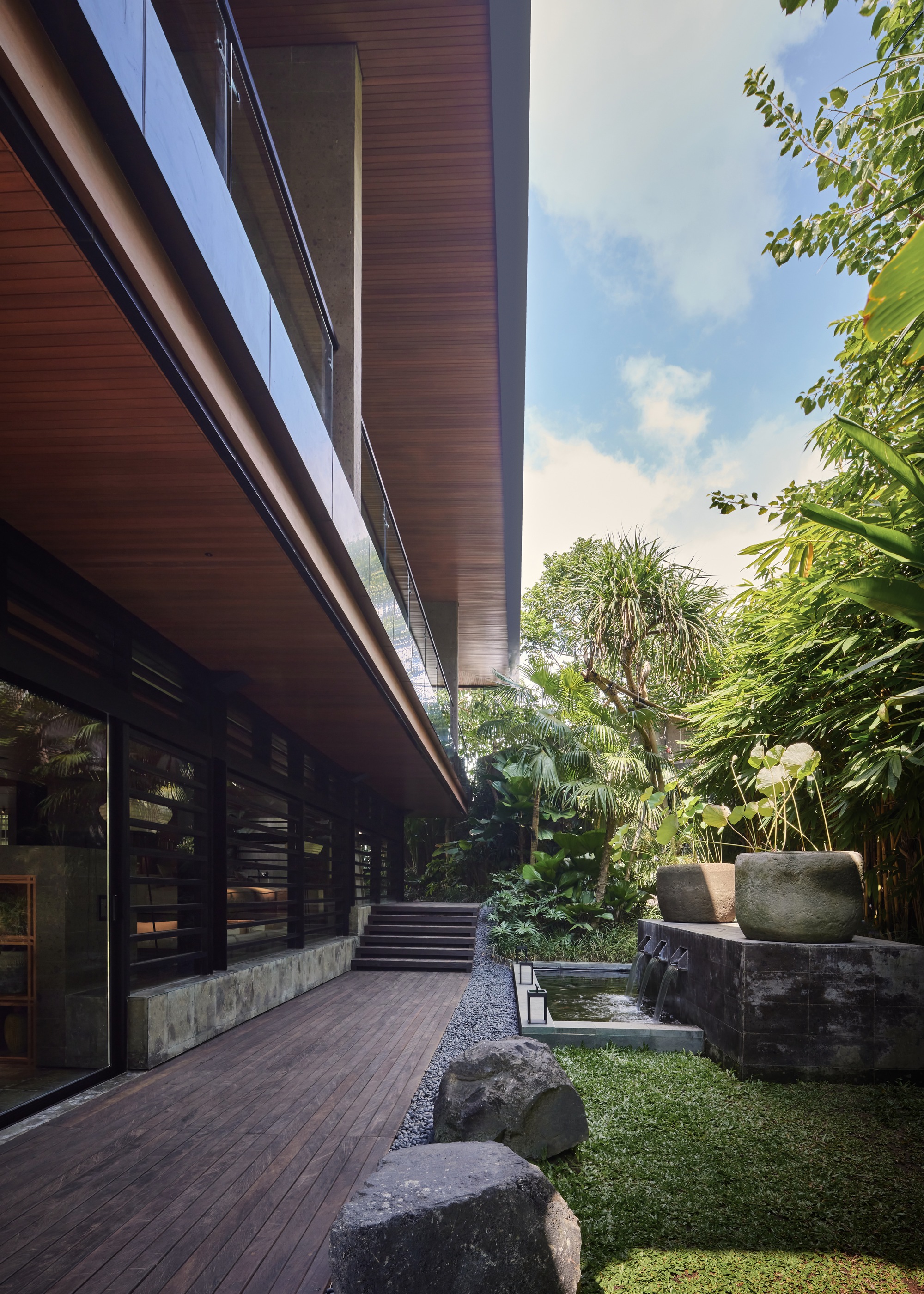 Timber deck running beneath a deep overhanging upper floor, with stone basin fountain, boulders and gravel edge under a partly cloudy sky