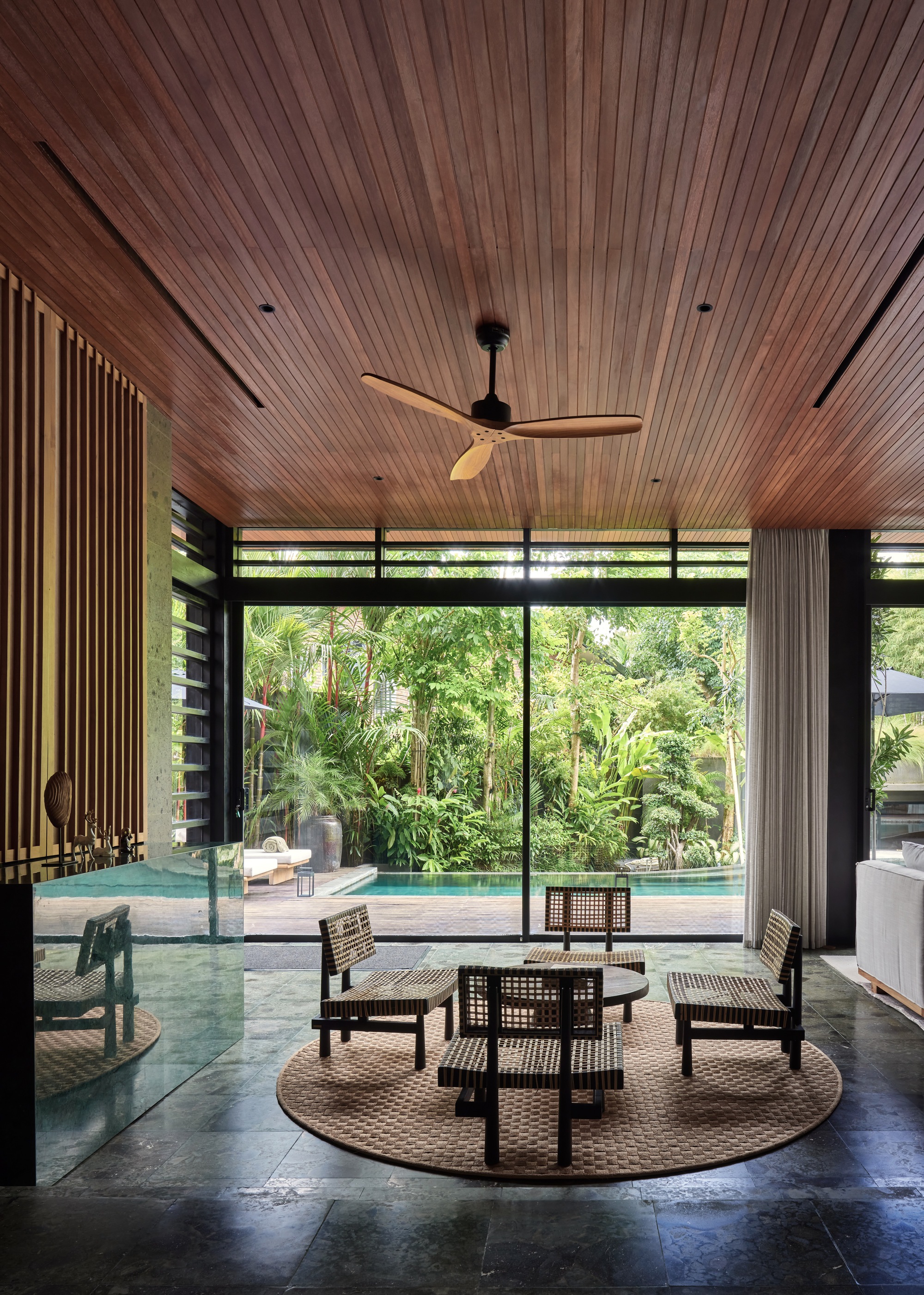 Living area with woven lounge chairs on a round rug, opening through glass doors to a pool and dense greenery in daylight