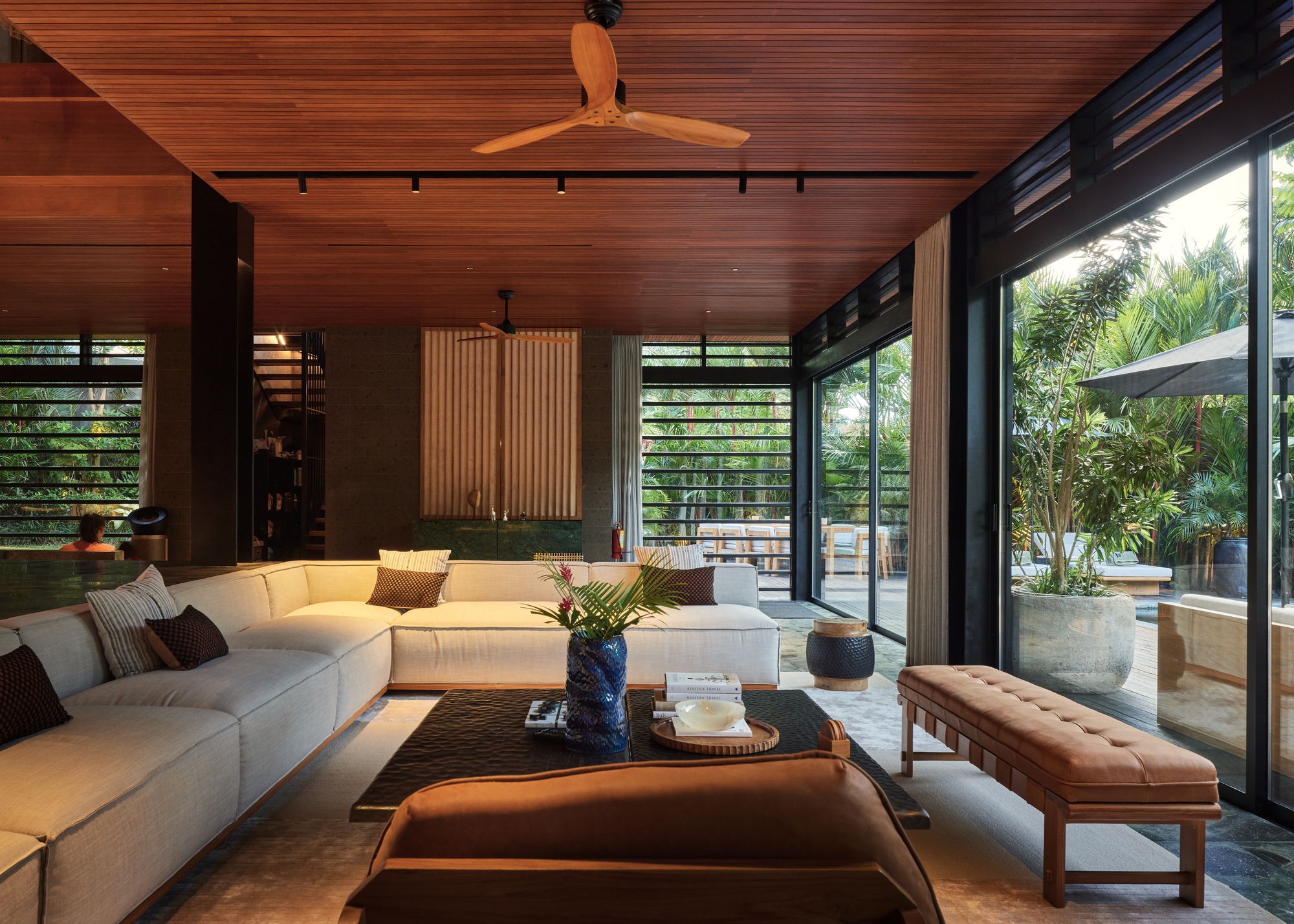 Sunken sofa, tufted leather bench and low table beneath a slatted timber ceiling, with sliding glass doors opening to a planted terrace