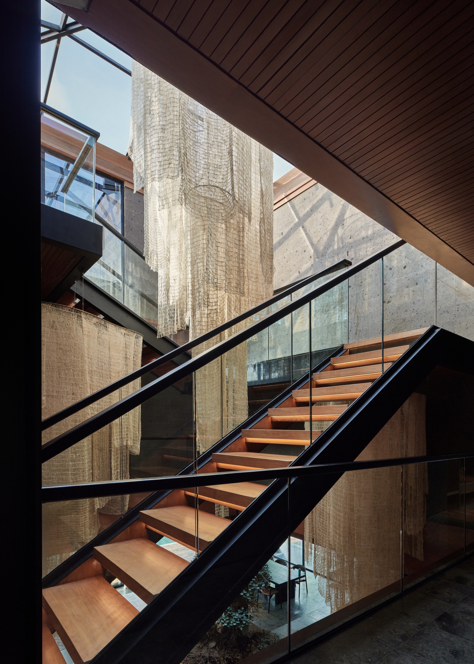 Open timber-tread stair with black steel stringers and glass guards beside a raw concrete wall and hanging metal mesh curtains, lit from above