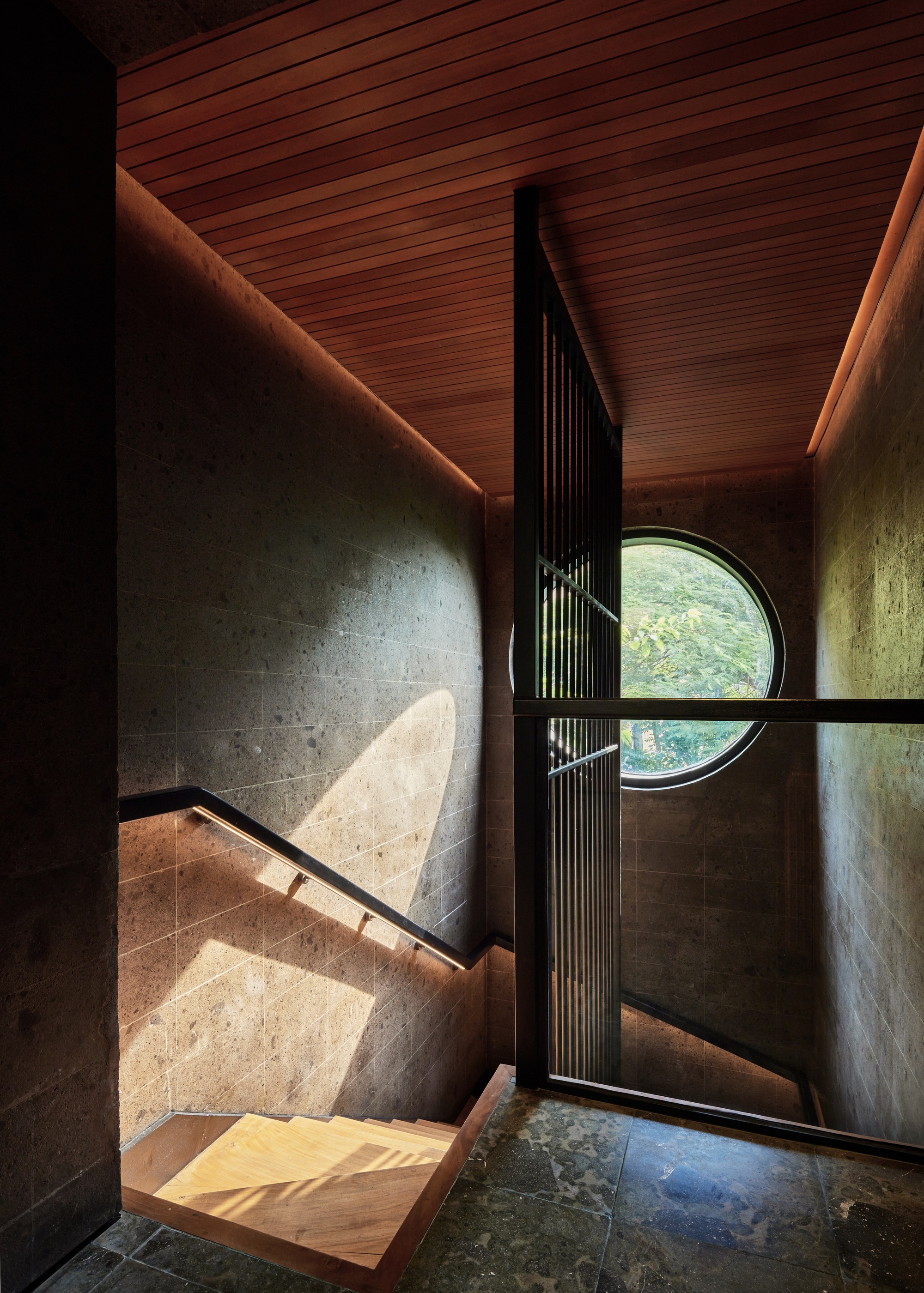 Stairwell landing with a round window, timber ceiling and stone walls, sunlight falling across the handrail and lower steps