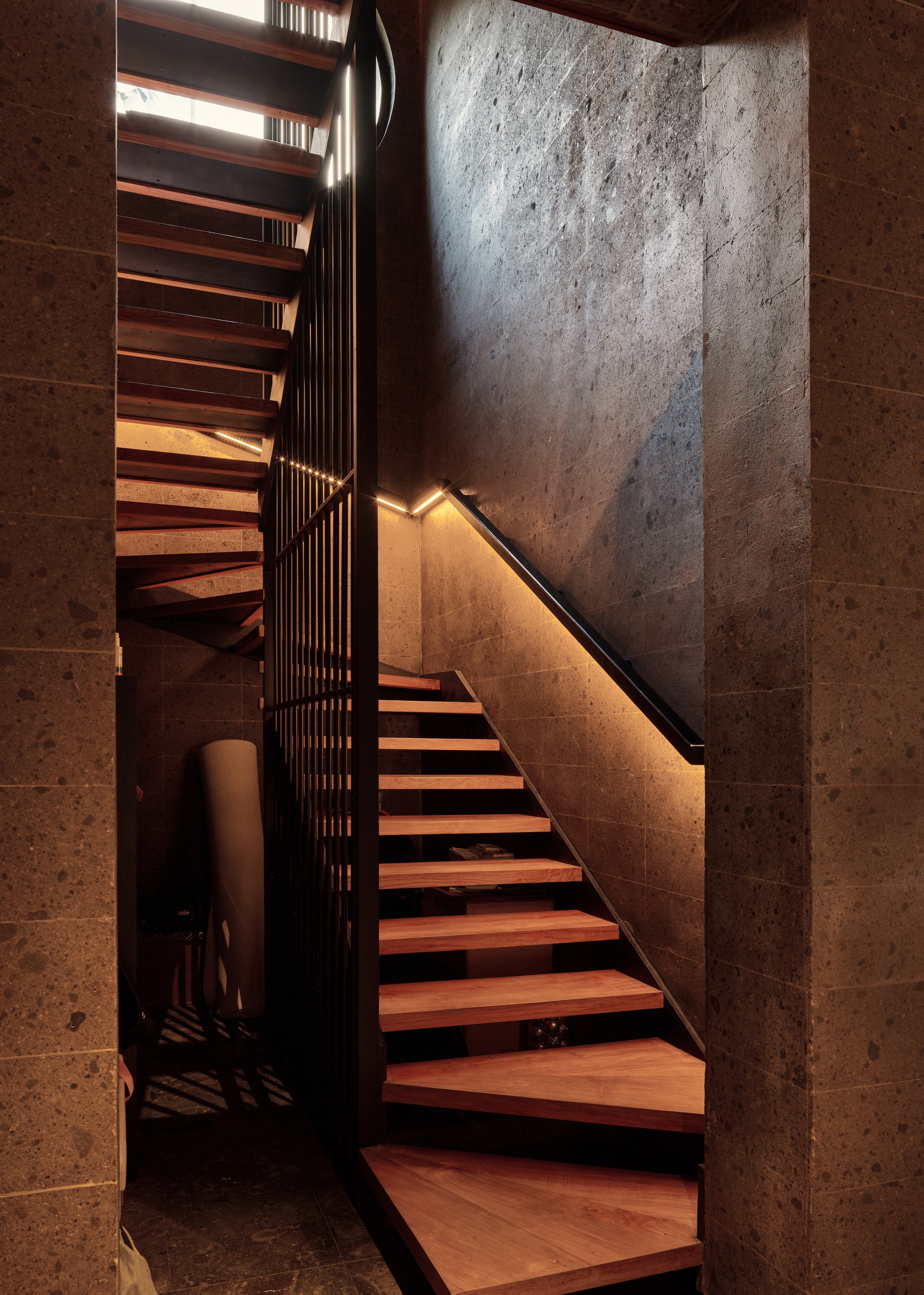 Timber stair with a lit handrail climbing between speckled stone walls and a vertical steel balustrade under a skylight