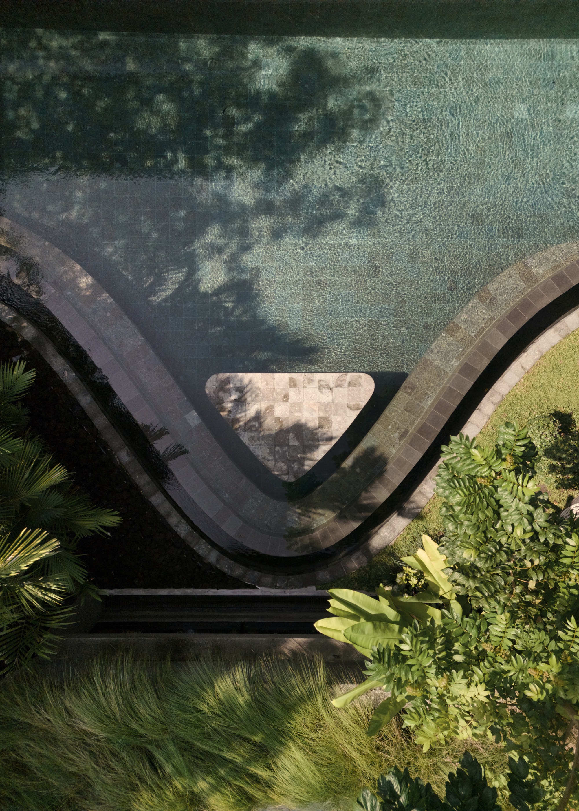 Top-down view of the wave-shaped pool edge with a triangular stone platform, tree shadows falling across the green tiled water