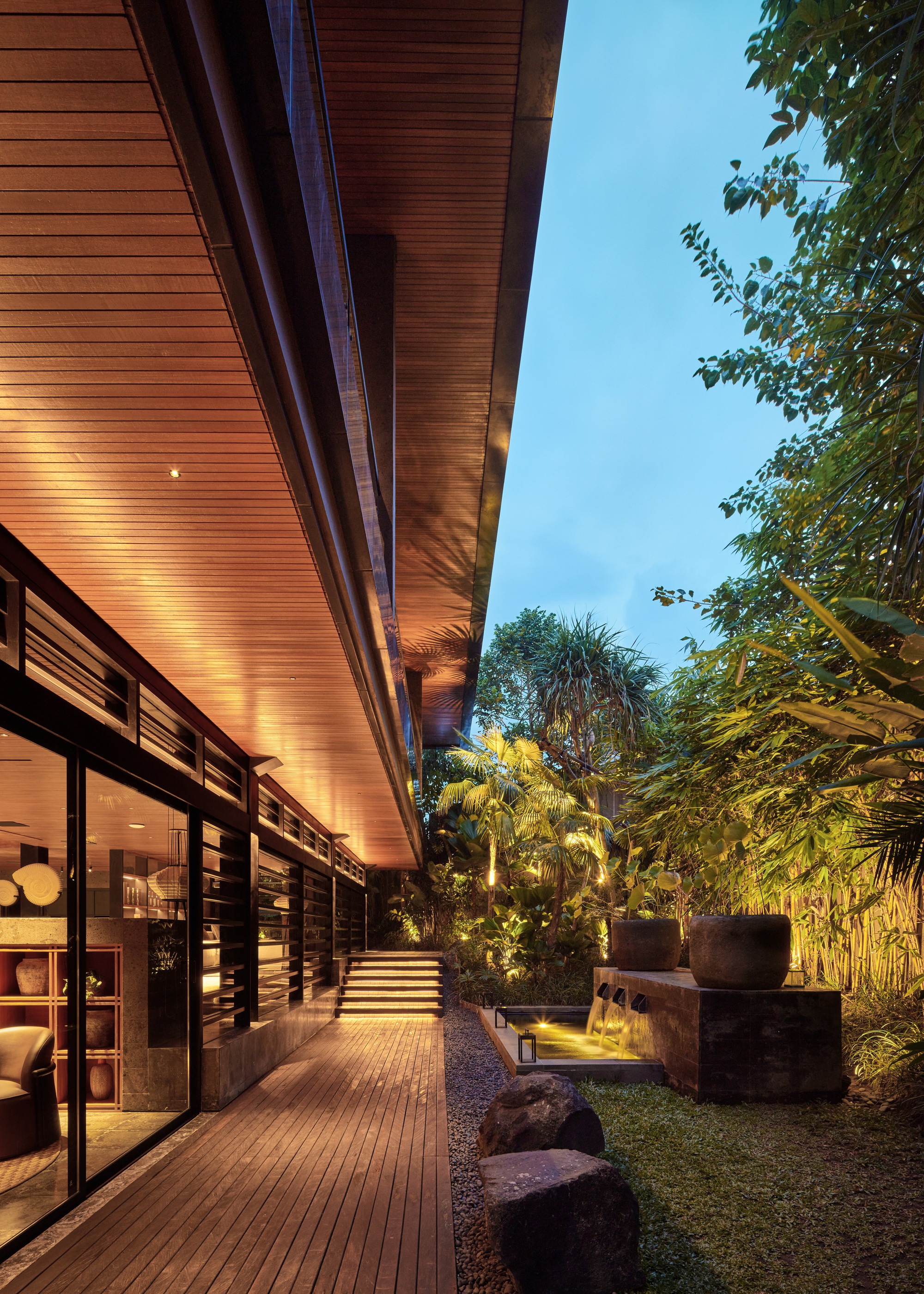 Timber-lined overhang above a wood deck with glass doors, stone steps and a small fountain, lit at dusk among palms