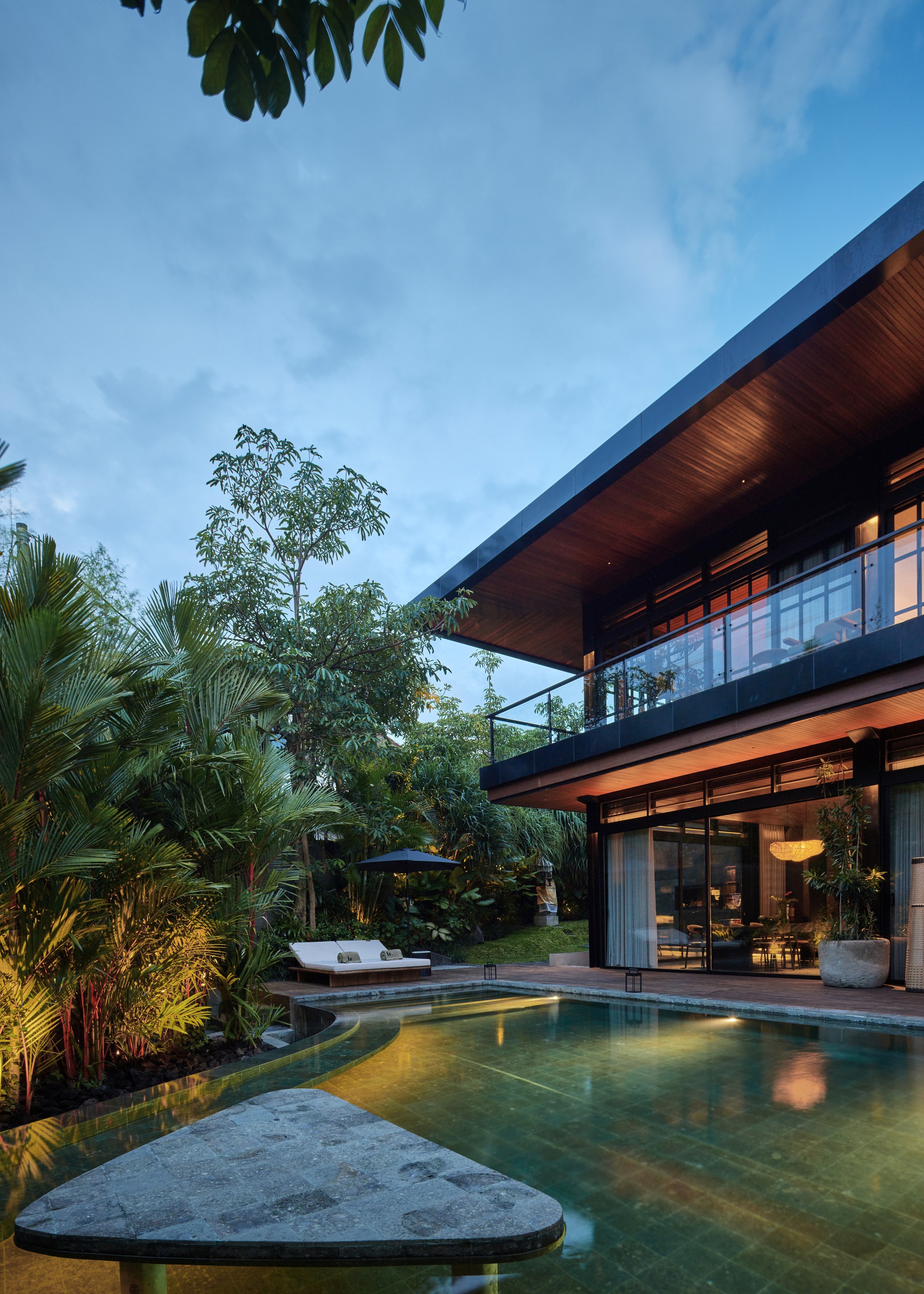 Pool edge with a stone slab in the foreground and the cantilevered upper floor with glass balustrade at blue hour