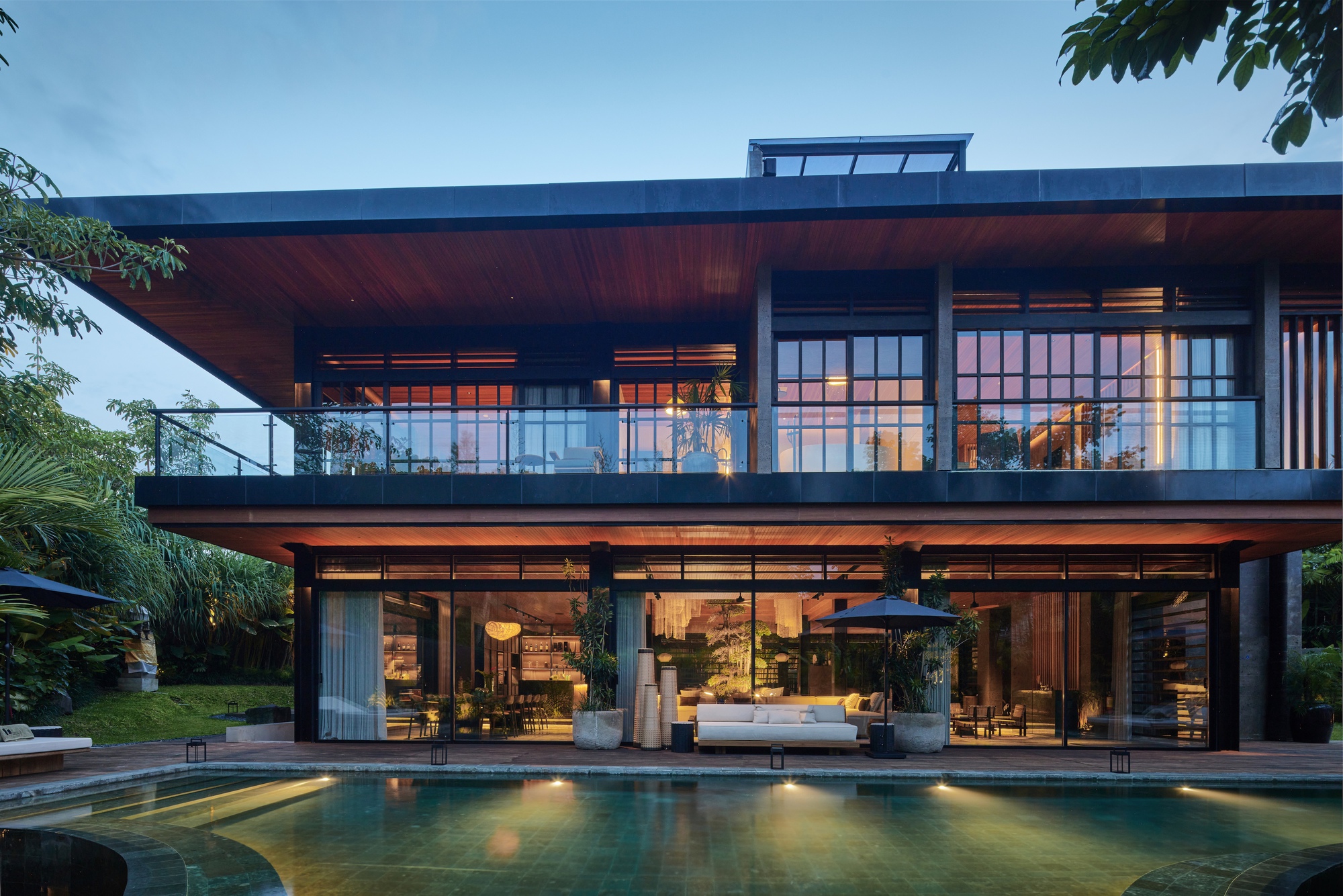 Two-storey pavilion facade with a wide timber-soffit roof, glazed living areas and a pool reflecting warm interior light at dusk