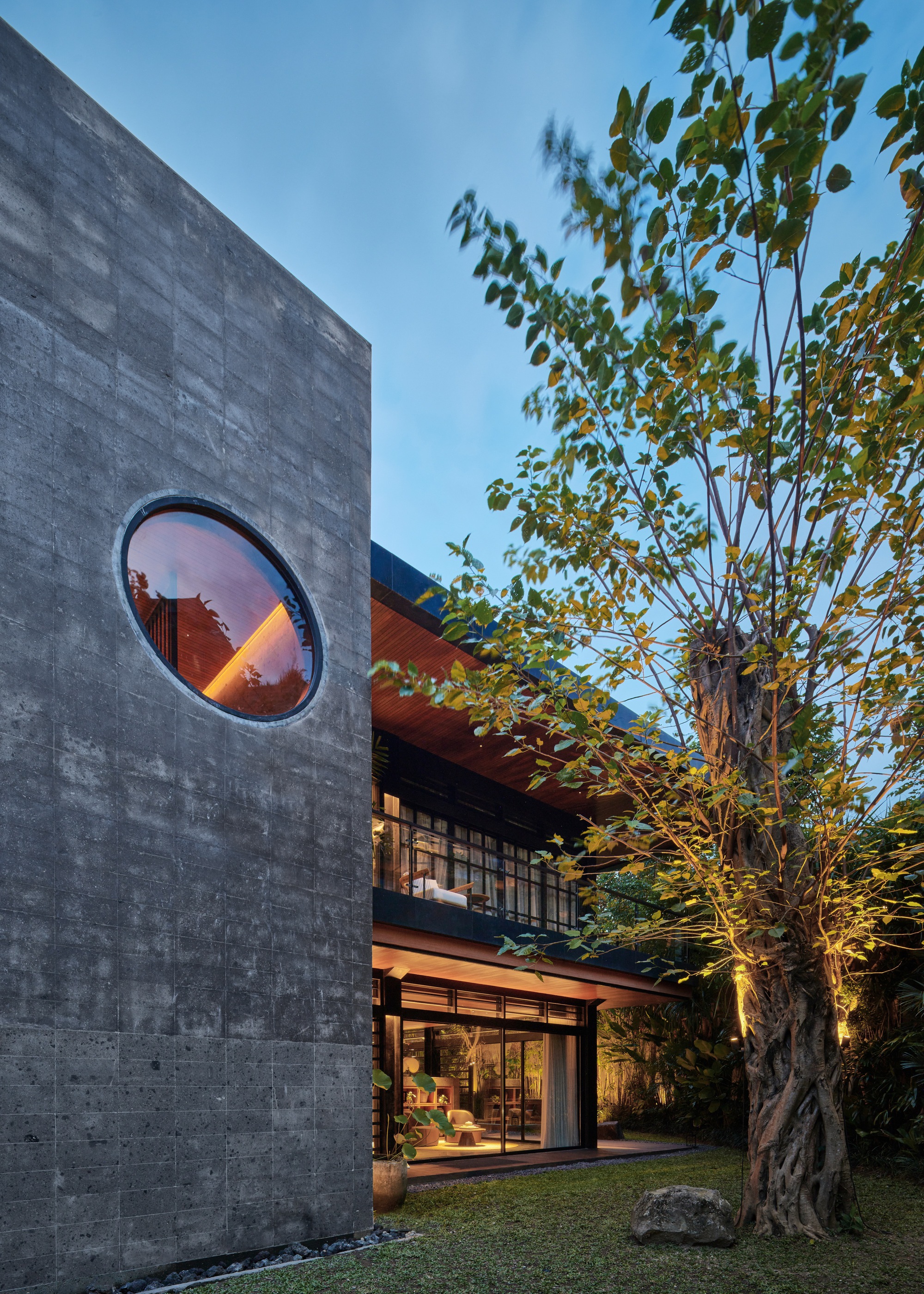 Board-marked concrete wall with a round window beside a lit tree trunk and the glazed house beyond at twilight