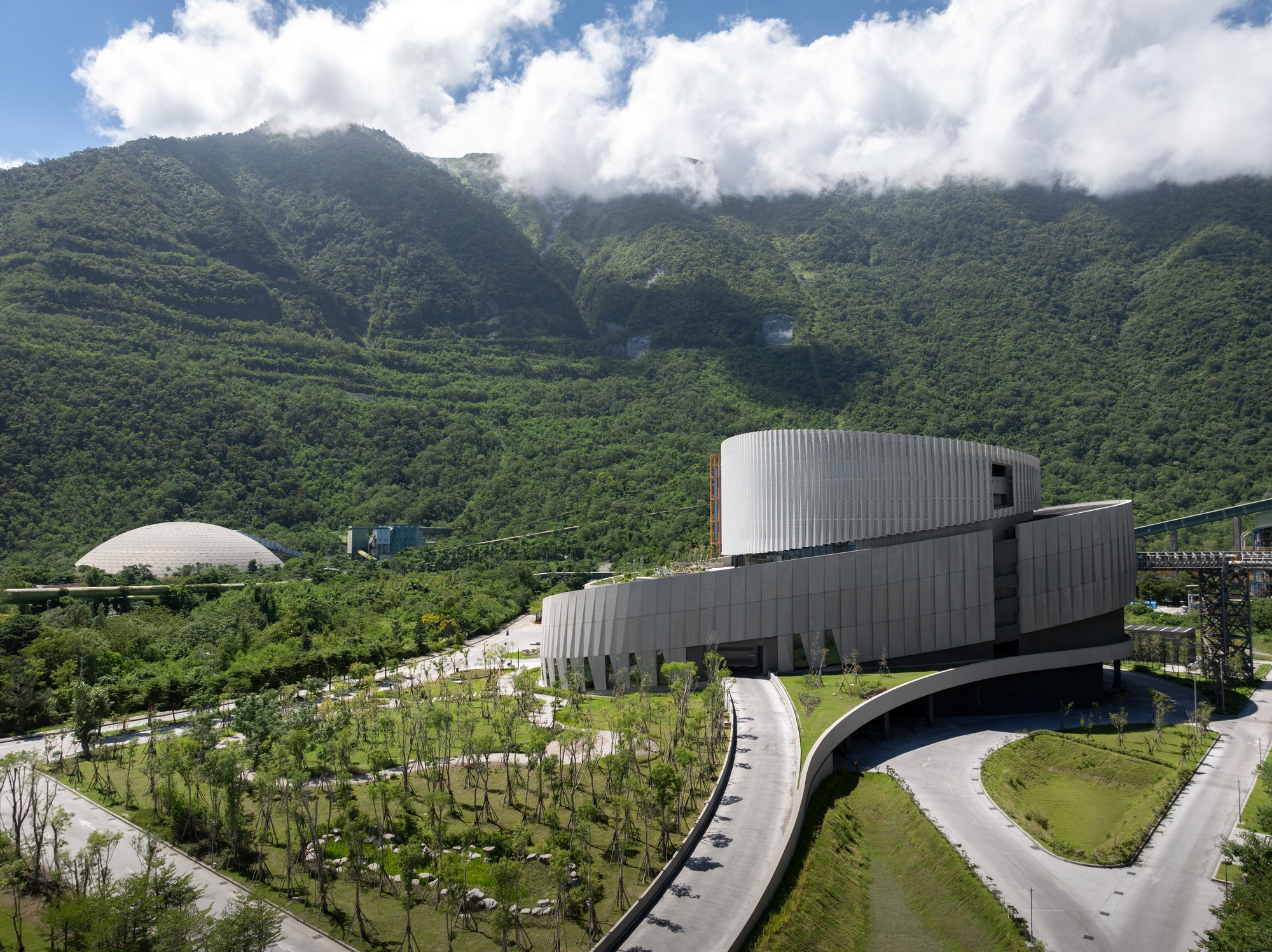 Aerial view of the round, pleated-metal building above a curving access road, with forested mountain and a white dome behind under clouds