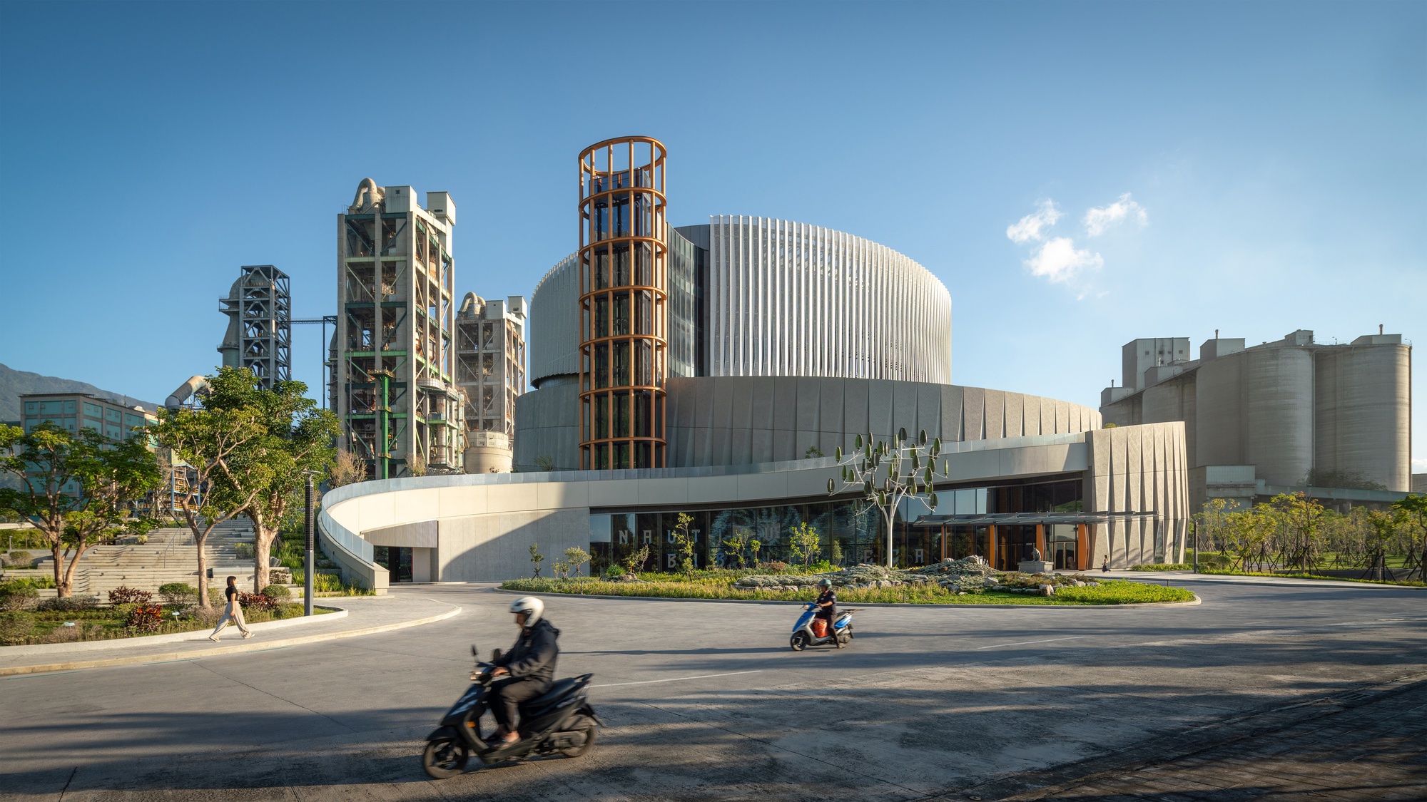 Street-level view of the cylindrical building with fluted upper drum and orange lattice tower, scooter riders passing in front of cement plant structures