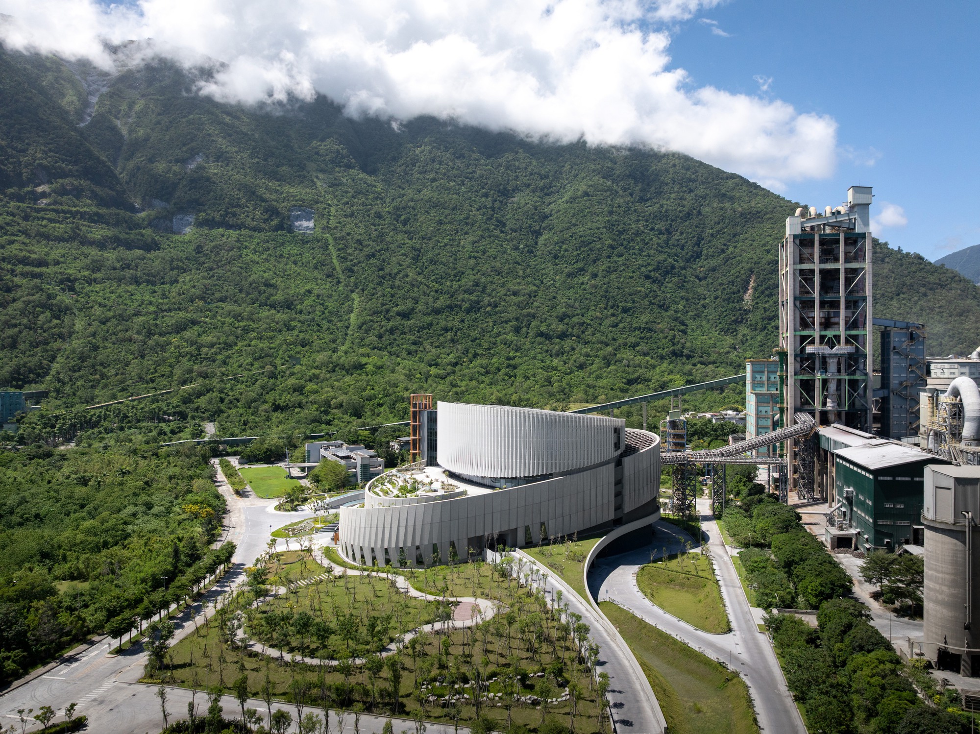Aerial view of the curved, ribbed facade beside cement plant towers and conveyors, with cloud-capped green mountainside rising behind