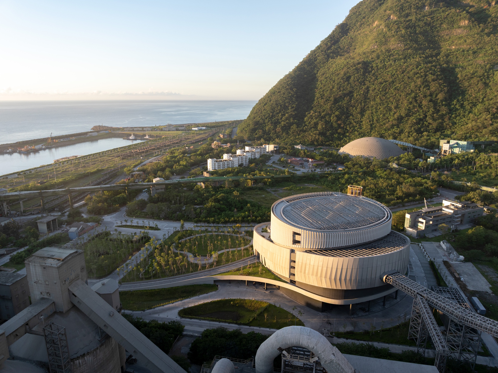 Aerial view of the drum-shaped roof with slatted top, a young tree grove, and the sea beyond in early morning light