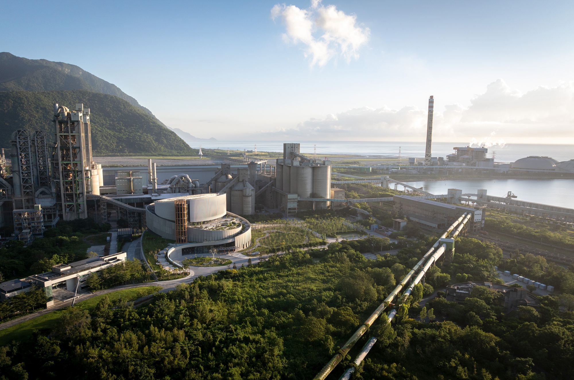 Distant aerial view of the round building among cement kilns, silos and a tall chimney, with pipe conveyors crossing dense green forest at dawn
