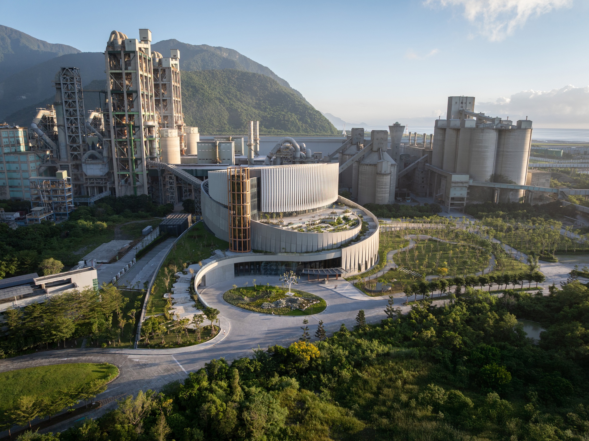 Aerial view of the terraced roof garden and orange lattice tower, with kiln towers and silos behind in low golden sunlight