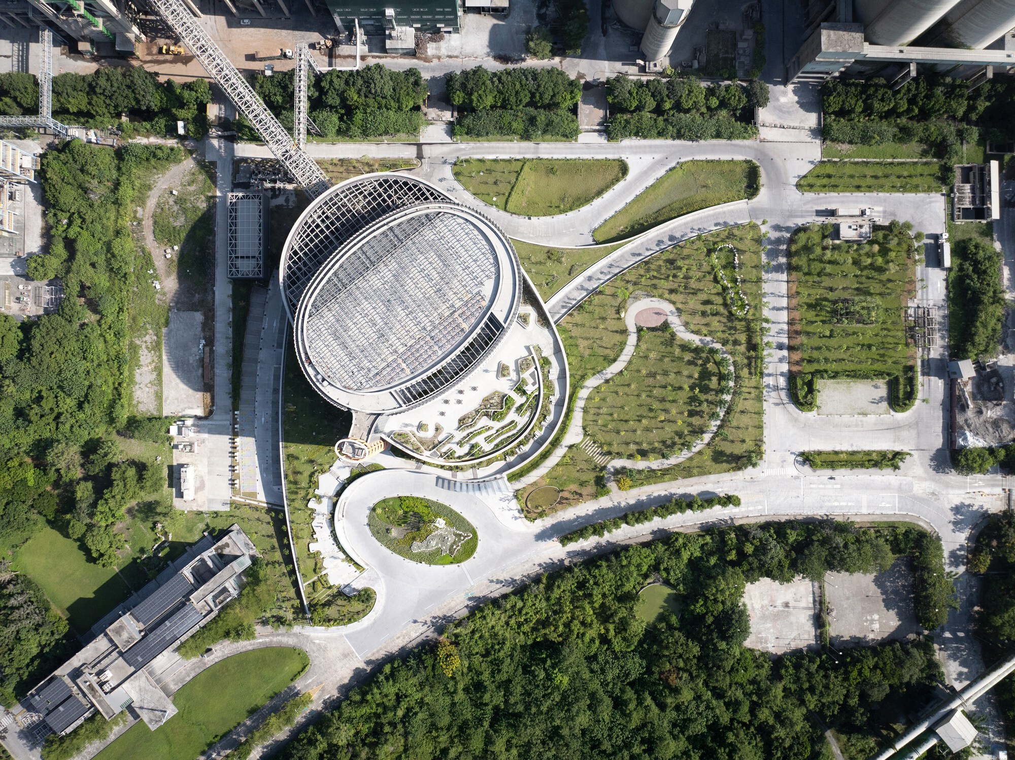 Top-down view of the oval glazed roof and curving paths and planted terraces, surrounded by dense green trees and industrial structures