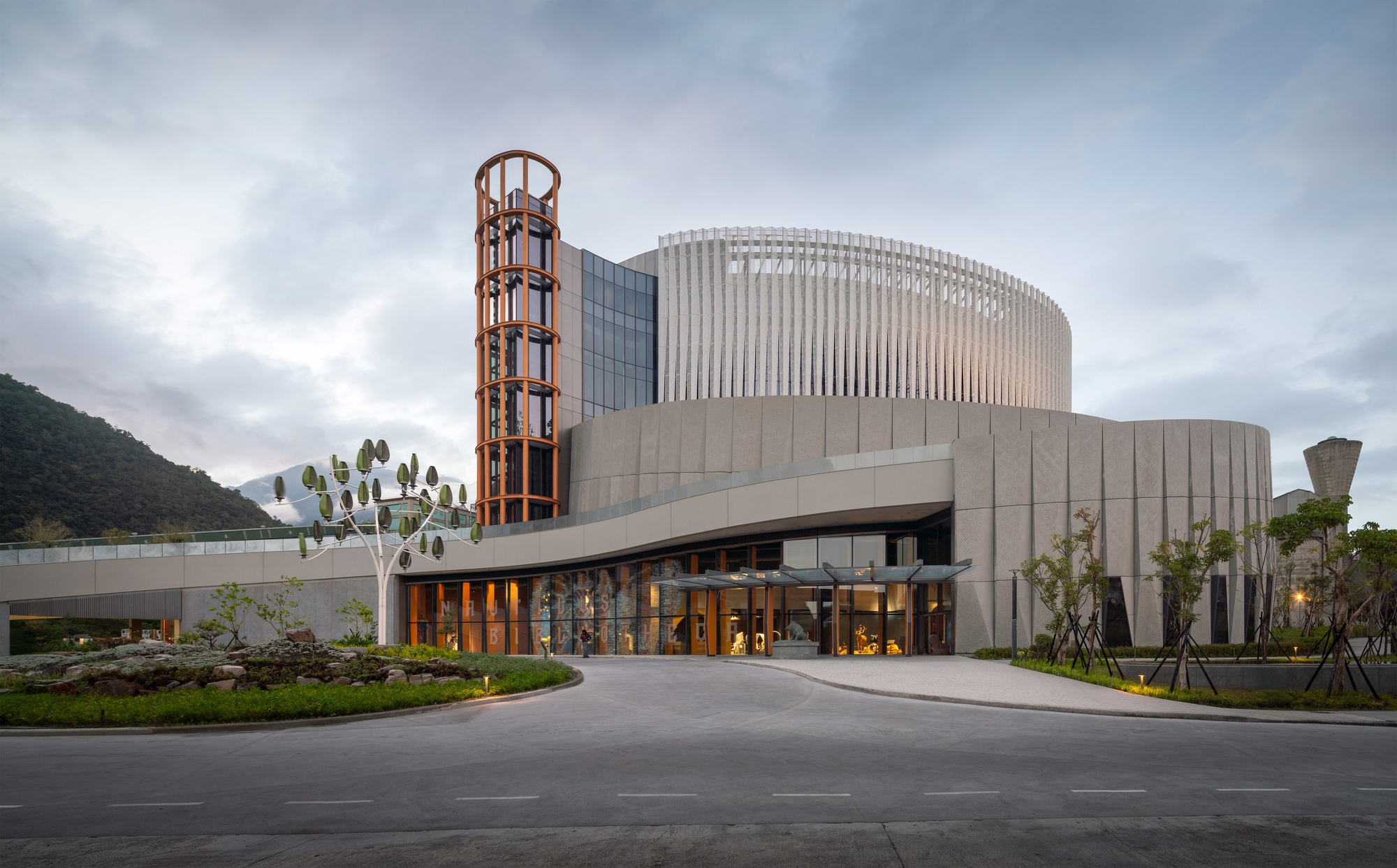 Entrance facade with a timber-framed glass tower and a curved perforated panel wall under a cloudy dusk sky