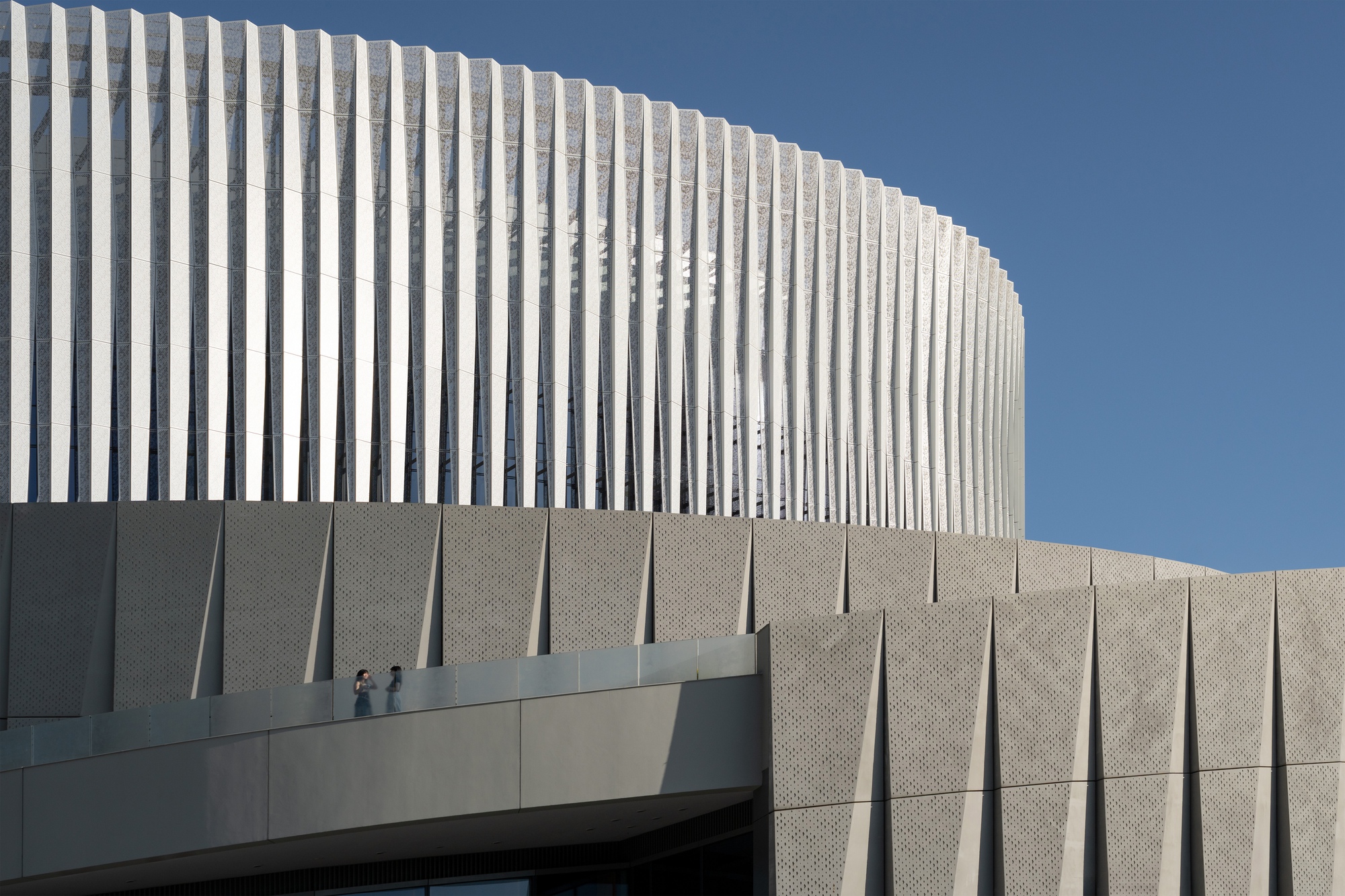 Close-up of vertical white fins above a faceted perforated panel wall, with two people on a balcony under clear blue sky