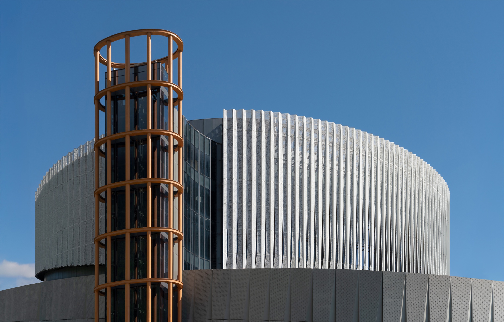 Orange steel cage tower beside the curved white fin facade, set against a deep blue sky