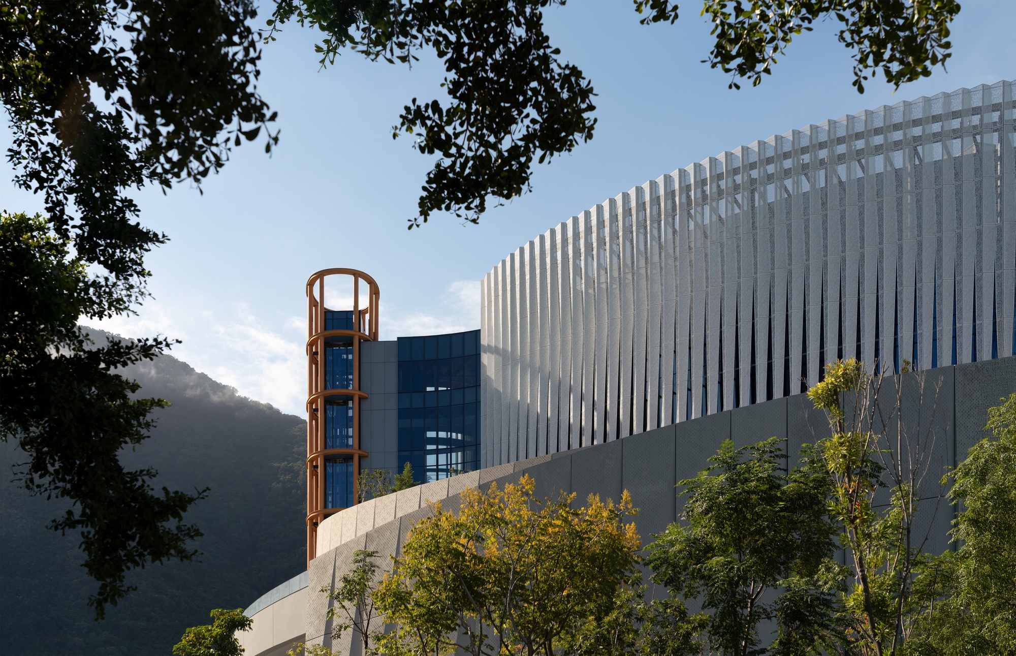 Fin-clad curved volume and orange tower seen through tree branches, with misty green hills in the afternoon light
