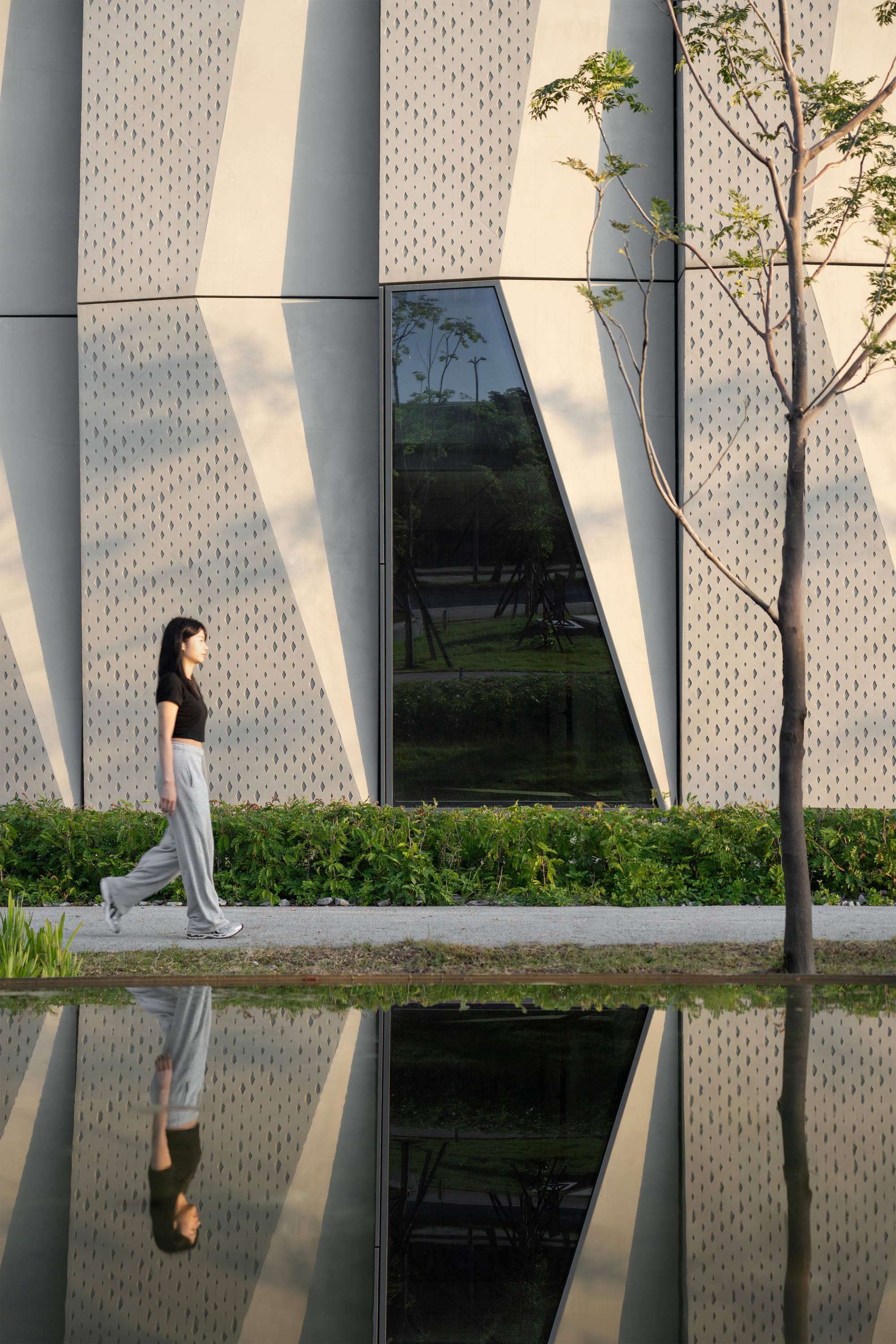 Woman walking past a faceted perforated panel wall and its reflecting pool, with a slim young tree in the foreground