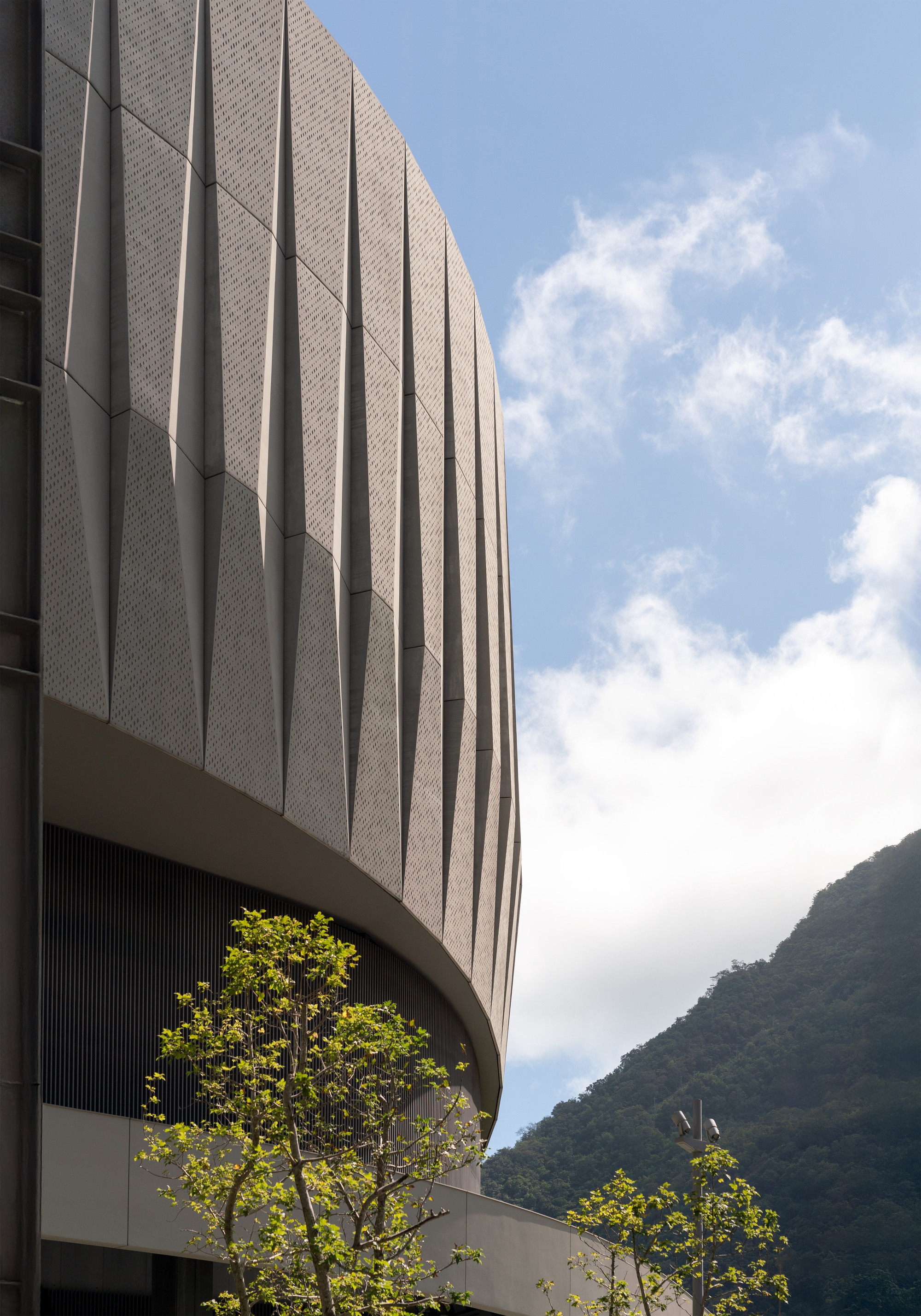 Curved facade of folded perforated panels rising above young trees, with a forested mountain slope behind under scattered clouds