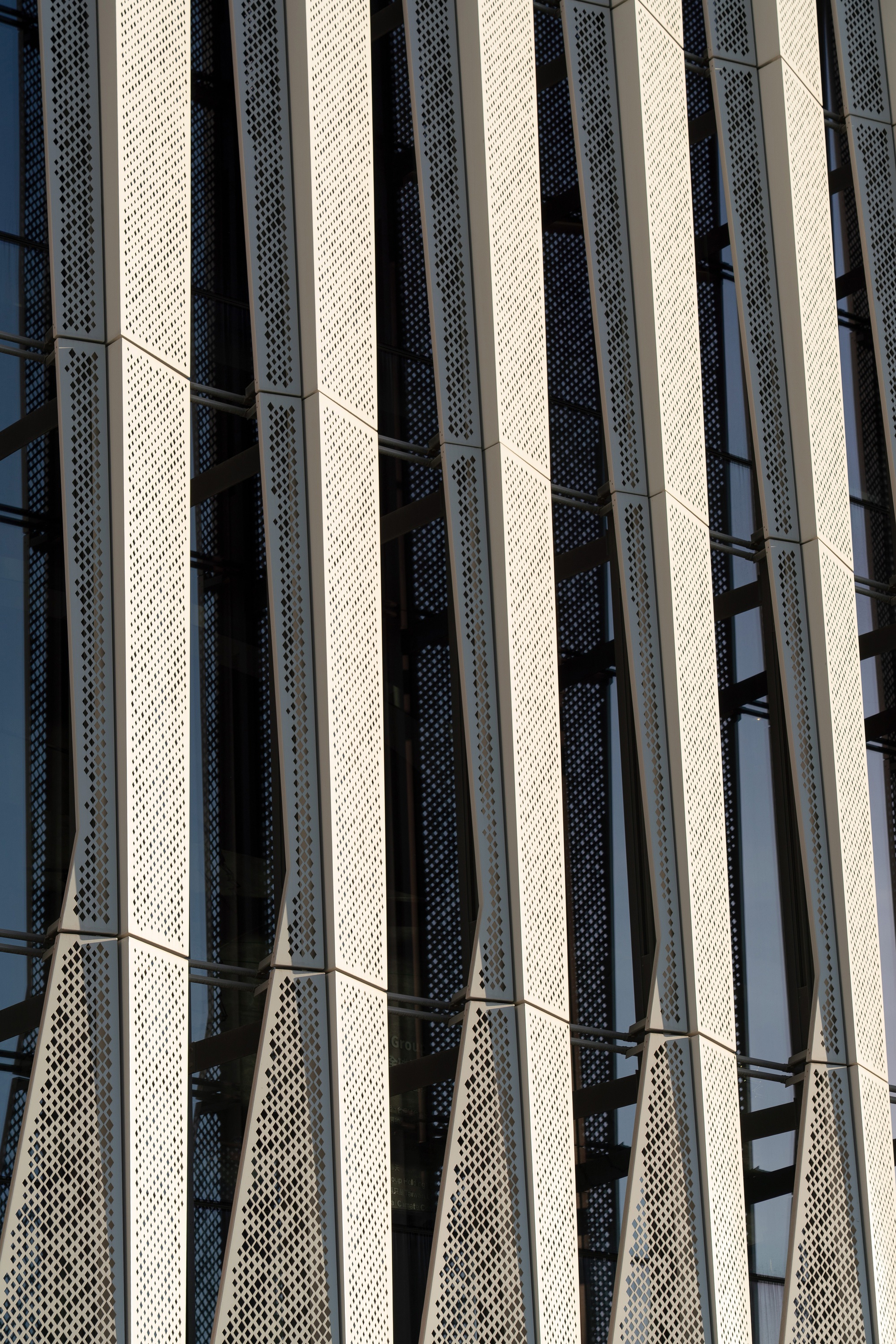 Close-up of vertical perforated metal fins twisting in front of glazing in low raking sunlight