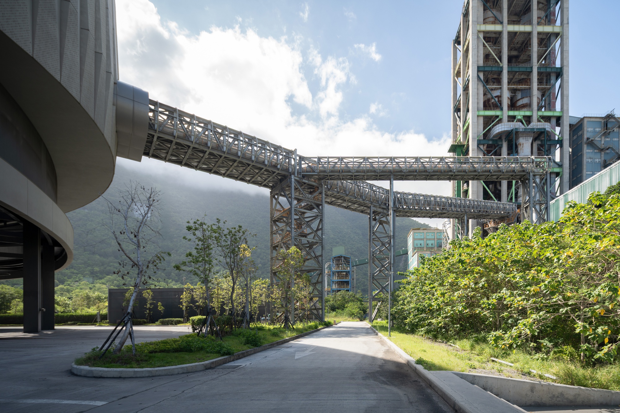 Steel truss conveyor bridge leaving the curved building and crossing a quiet road toward an industrial tower, with mist on the hills