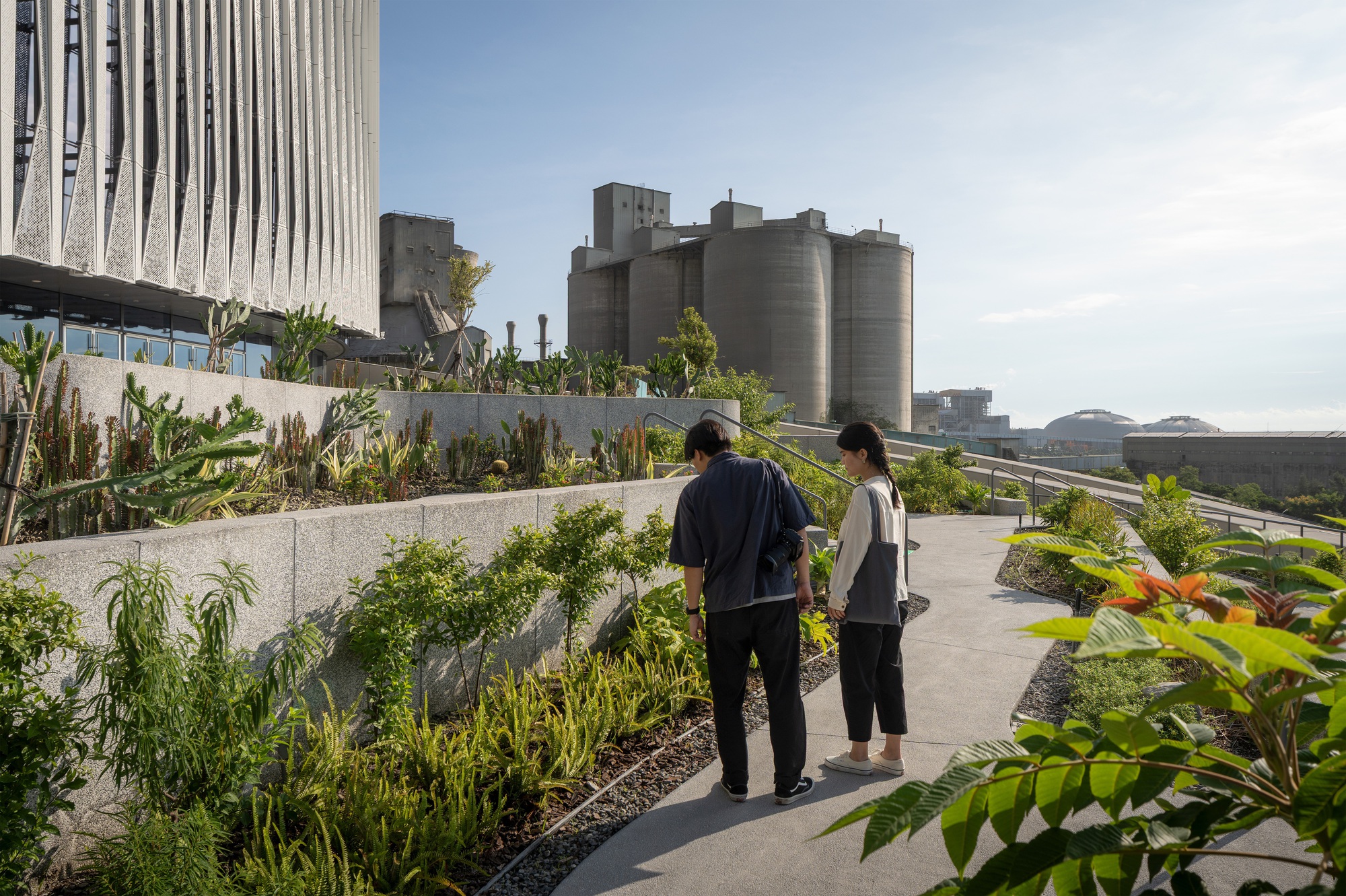Two visitors on a rooftop garden path lined with planted beds, with cement silos rising in the distance at golden hour