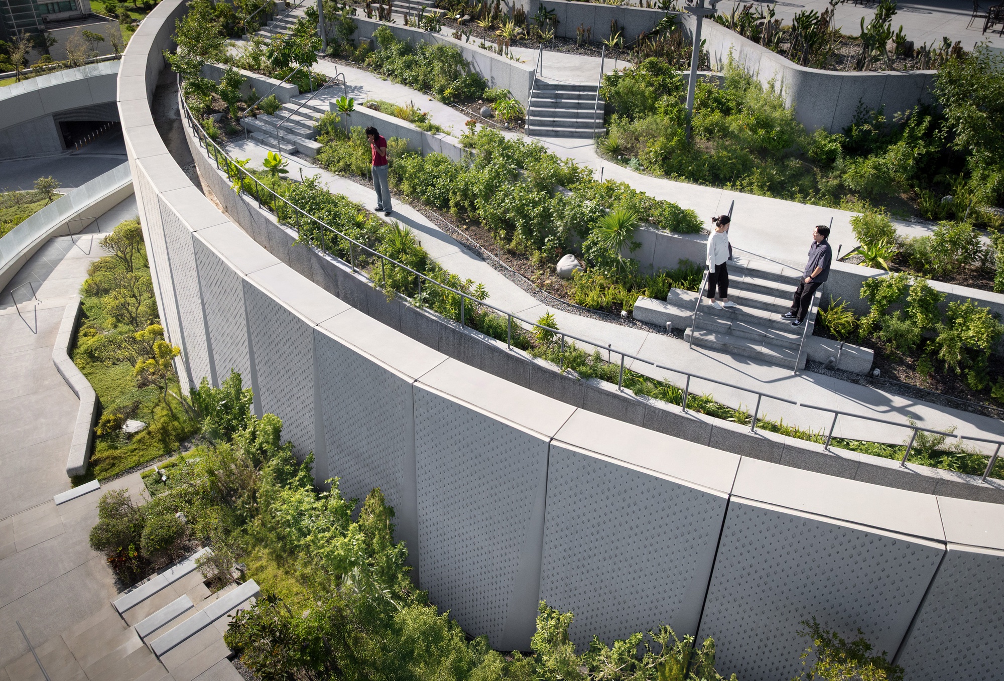 Looking down on a sweeping perforated concrete wall above stepped garden terraces where three people stand among shrubs in daylight