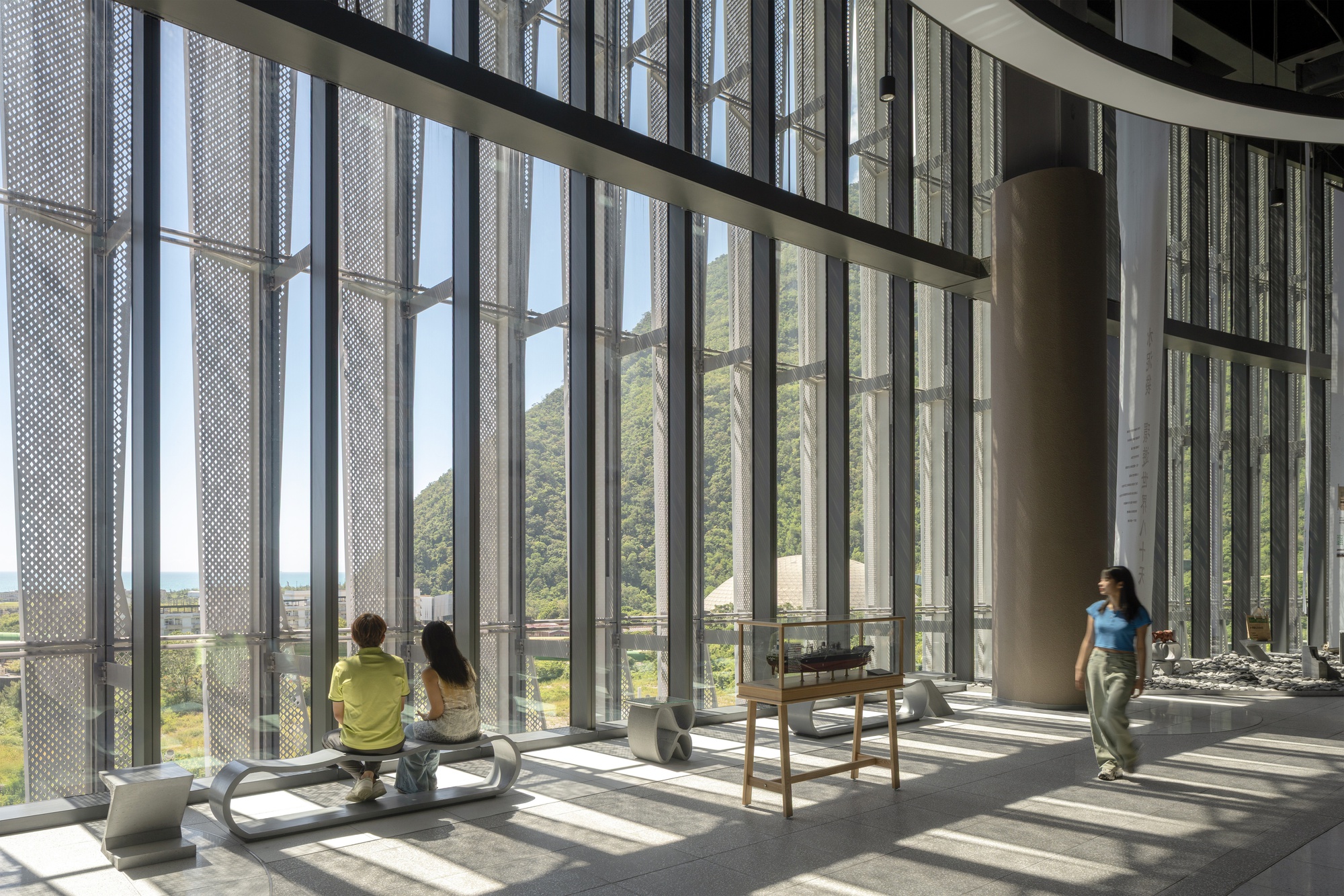 Double-height glazed hall with hanging perforated screens, a display case and visitors on benches, with forested hills beyond in sunlight
