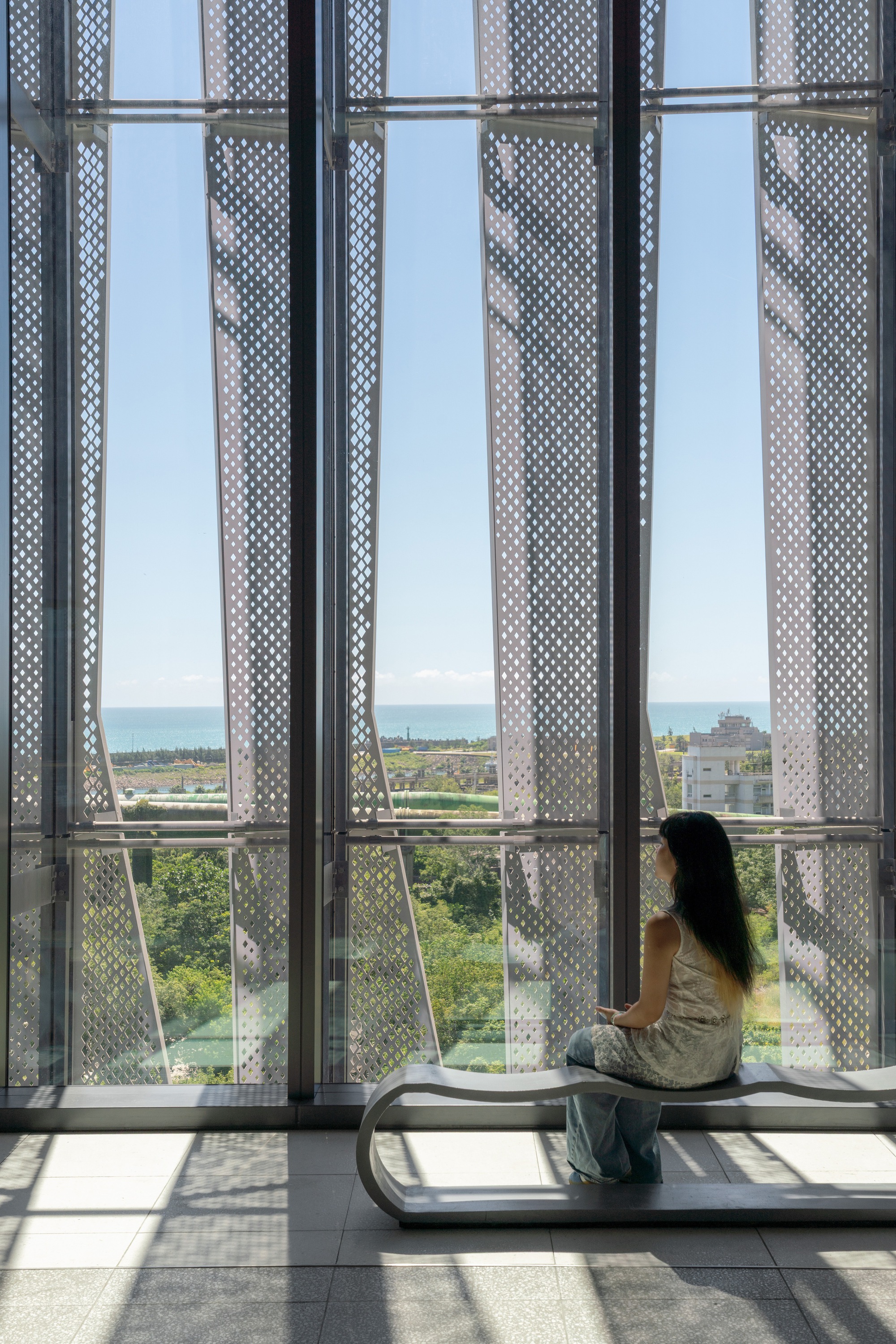 Visitor seated on a curved steel bench before tall glass and perforated screens, with the sea on the horizon