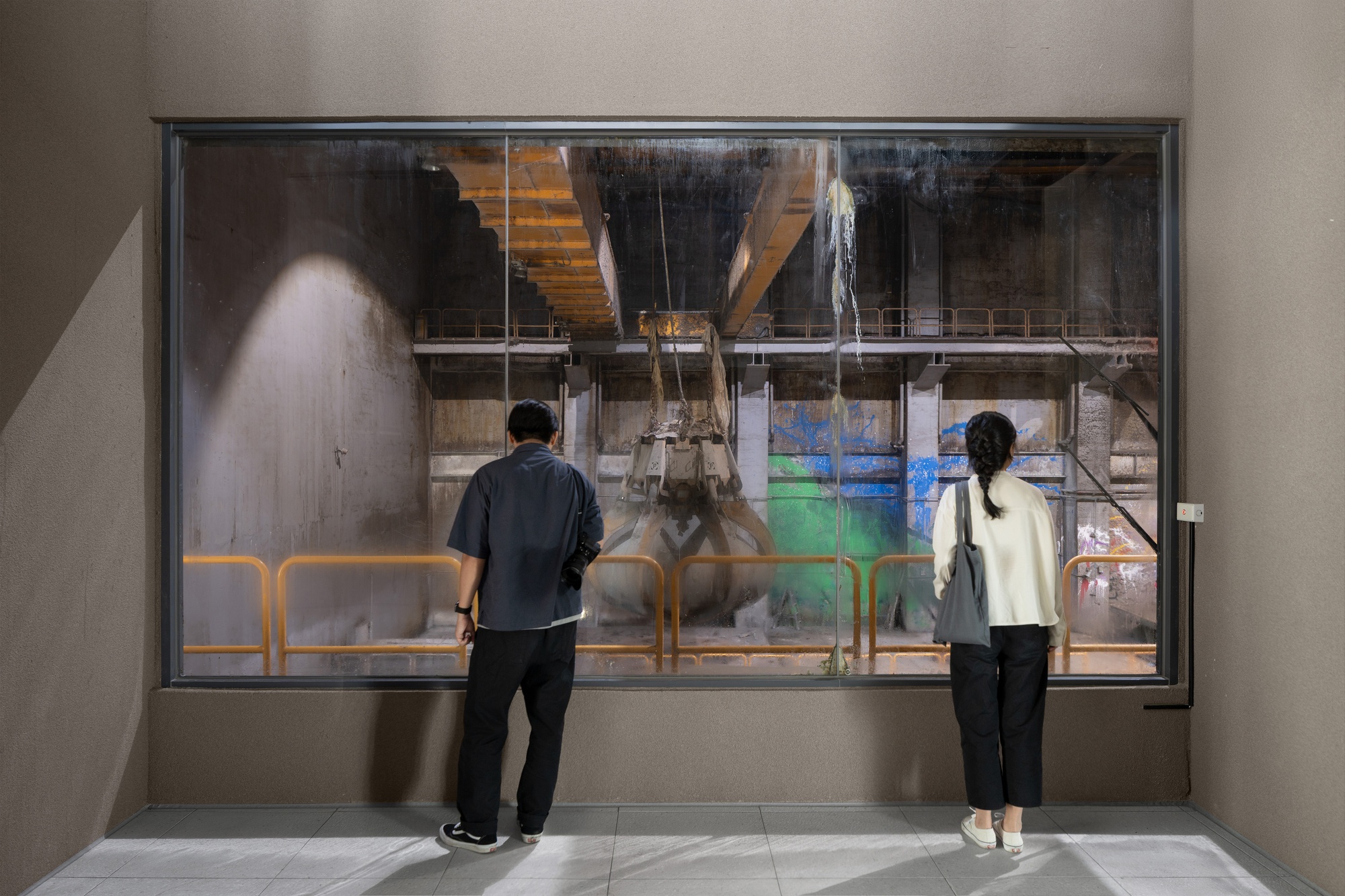 Two visitors at a large interior window facing a concrete pit with a hanging grab bucket and orange railings