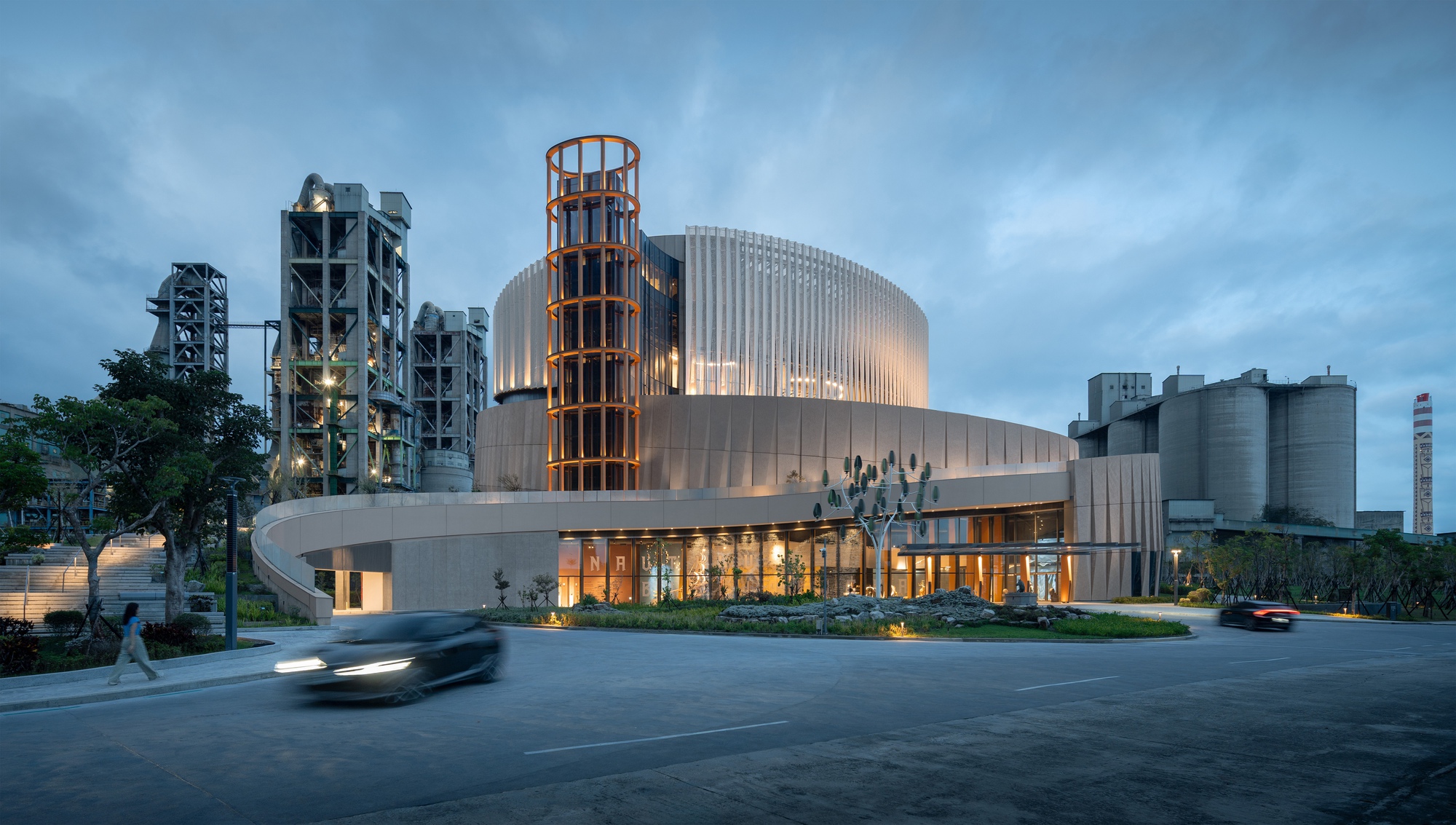 Corner view of the drum-shaped building with an orange lattice tower, cement plant structures behind and a car passing at dusk